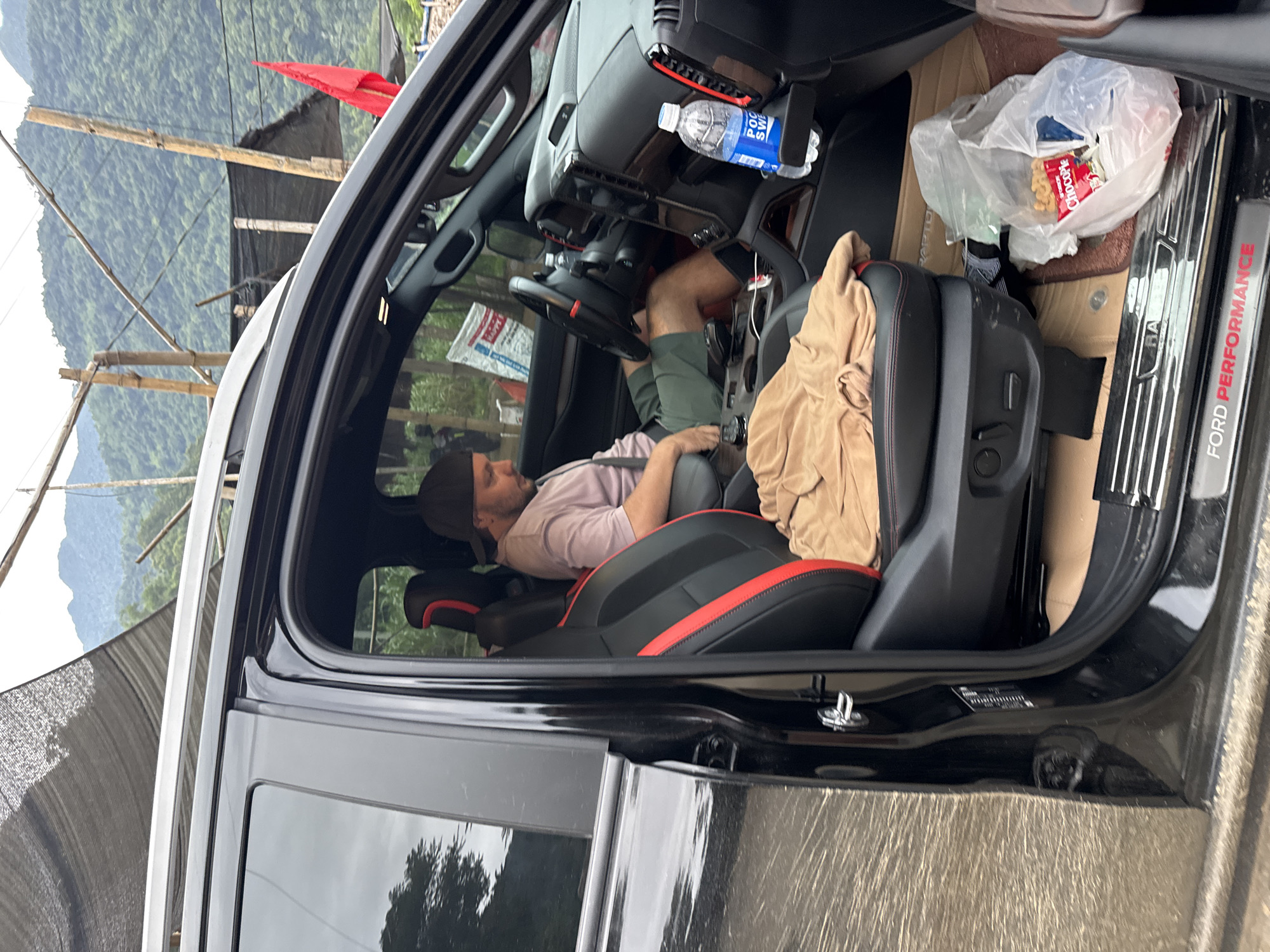 A man is seated in the driver's seat of a black Ford Raptor vehicle in Cổ Lũng Commune, Vietnam. The man is wearing a light pink t-shirt and green shorts. He is wearing a black baseball cap. The vehicle's interior is visible, with black leather seats accented with red stitching. A beige cloth is draped over the passenger seat. A plastic bag containing snacks is on the floor of the passenger side. A bottle of Pocari Sweat water is in the cup holder. Outside the vehicle, mountains and bamboo structures are visible. The ground is muddy.