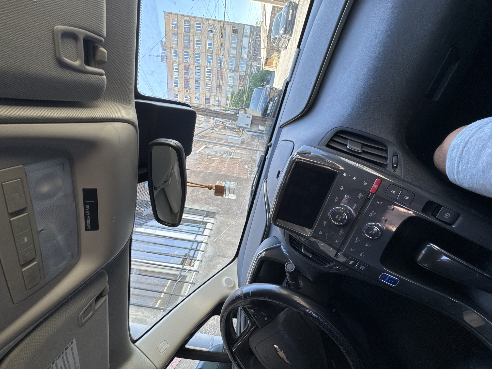 The image is an interior shot of a car, taken from the driver's perspective, with a view through the windshield of a building in Yerevan, Armenia. The building is an old, multi-story structure with numerous windows and what appears to be a densely populated residential neighborhood. Below the building, there's a courtyard or street area with various utility lines and what seems to be a lattice or grate structure.
Inside the car, the dashboard, steering wheel, and front passenger seat are visible. The car's infotainment system and controls are visible. A small air freshener hangs from the rearview mirror. On the passenger side, part of an arm or sleeve of a person is visible. The interior of the car appears to be modern, with a dark-colored dashboard and steering wheel. The car is situated on a street or road, possibly with other vehicles visible in the background. The overall scene suggests an urban environment.
