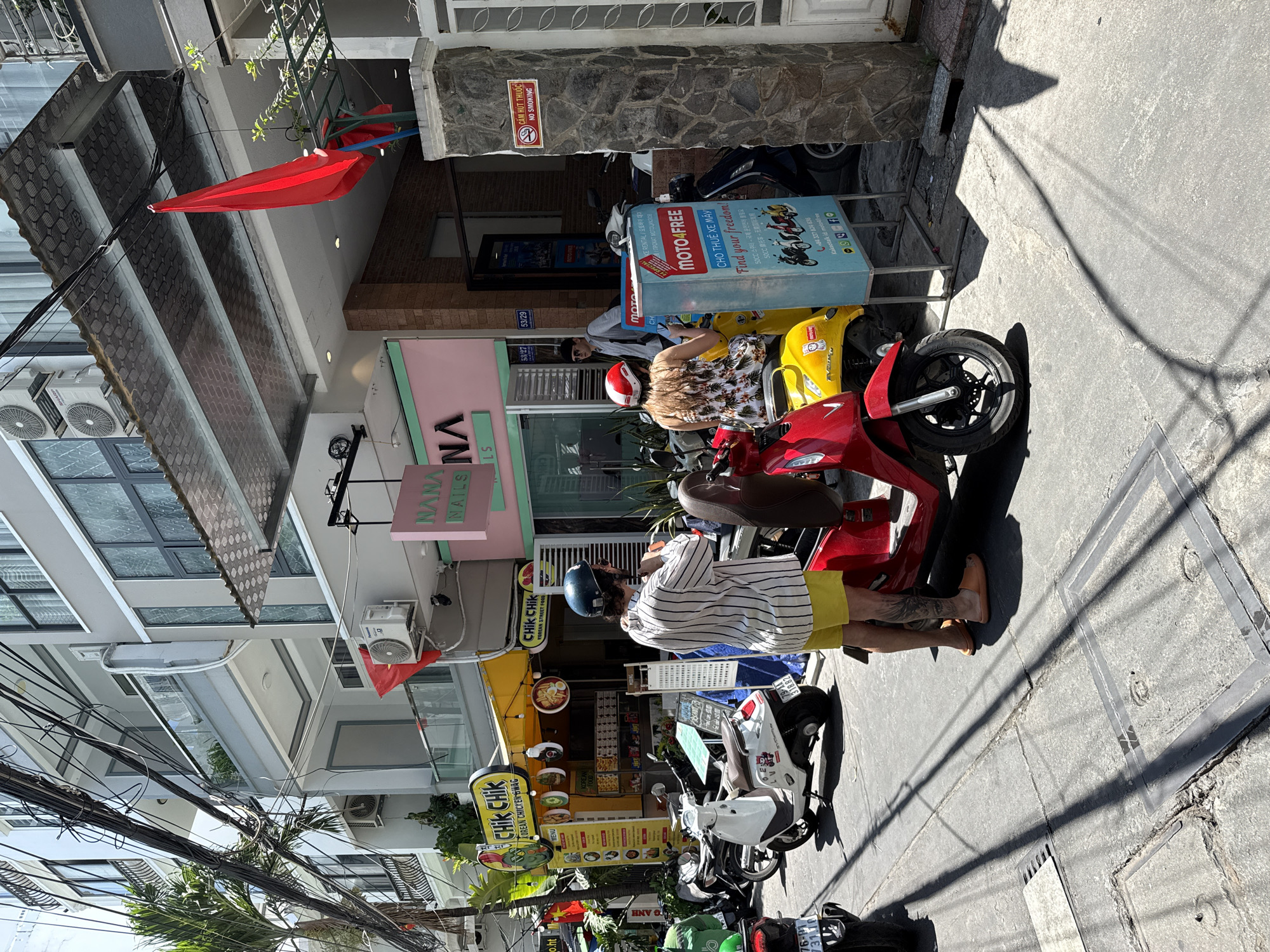 The image is taken from a street-level perspective, possibly in Nha Trang Ward, Vietnam, based on the visible businesses and signage. Two individuals are sitting on parked scooters. One scooter is red and the other is yellow. Both people appear to be looking at a blue advertising sign for a scooter rental service called "MOTO4FREE." The sign features various images of scooters and contact information. Further down the street, there are more parked scooters in white, green, and black.
Several businesses are visible in the background. These include a nail salon called "NANA NAILS" and a Korean street food restaurant called "CHIK CHIK." A "NO SMOKING" sign is attached to the stone facade of the upper portion of a building. Red flags are draped from the buildings. Above, many power cables can be seen.
The image is taken in daylight, with shadows cast on the street. The environment suggests a busy urban scene.