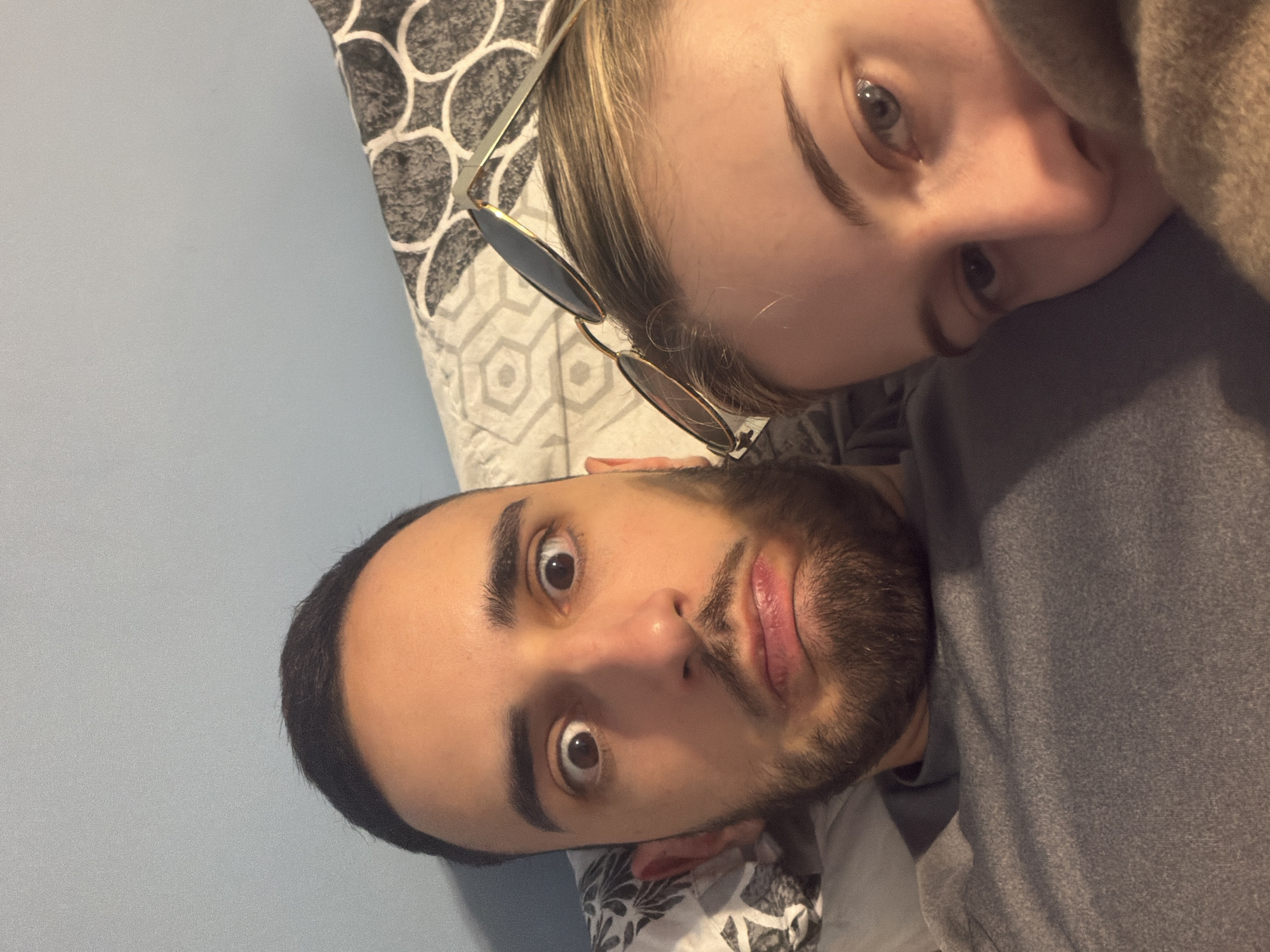 The scene depicts two individuals indoors, likely in a bedroom setting. A male subject occupies the left and central portion of the frame, positioned as if lying down. He has dark hair and a beard, and is seen looking directly at the camera with wide eyes and a pursed, slightly downturned mouth, indicating an exaggerated or playful expression. He wears a grey t-shirt. To the right, partially visible and adjacent to the male, is a female subject. Her right eye, part of her nose, and blonde hair are discernible. Gold-framed sunglasses with dark lenses rest on top of her head. A brown or tan textured fabric partially covers her lower face. Both individuals appear to be resting on pillows with a white and dark grey geometric pattern. A plain light blue wall is visible in the background. The image is a close-up, seemingly a selfie. No discernible features or landmarks within the image allow for the identification of a specific location within Plovdiv, Bulgaria.