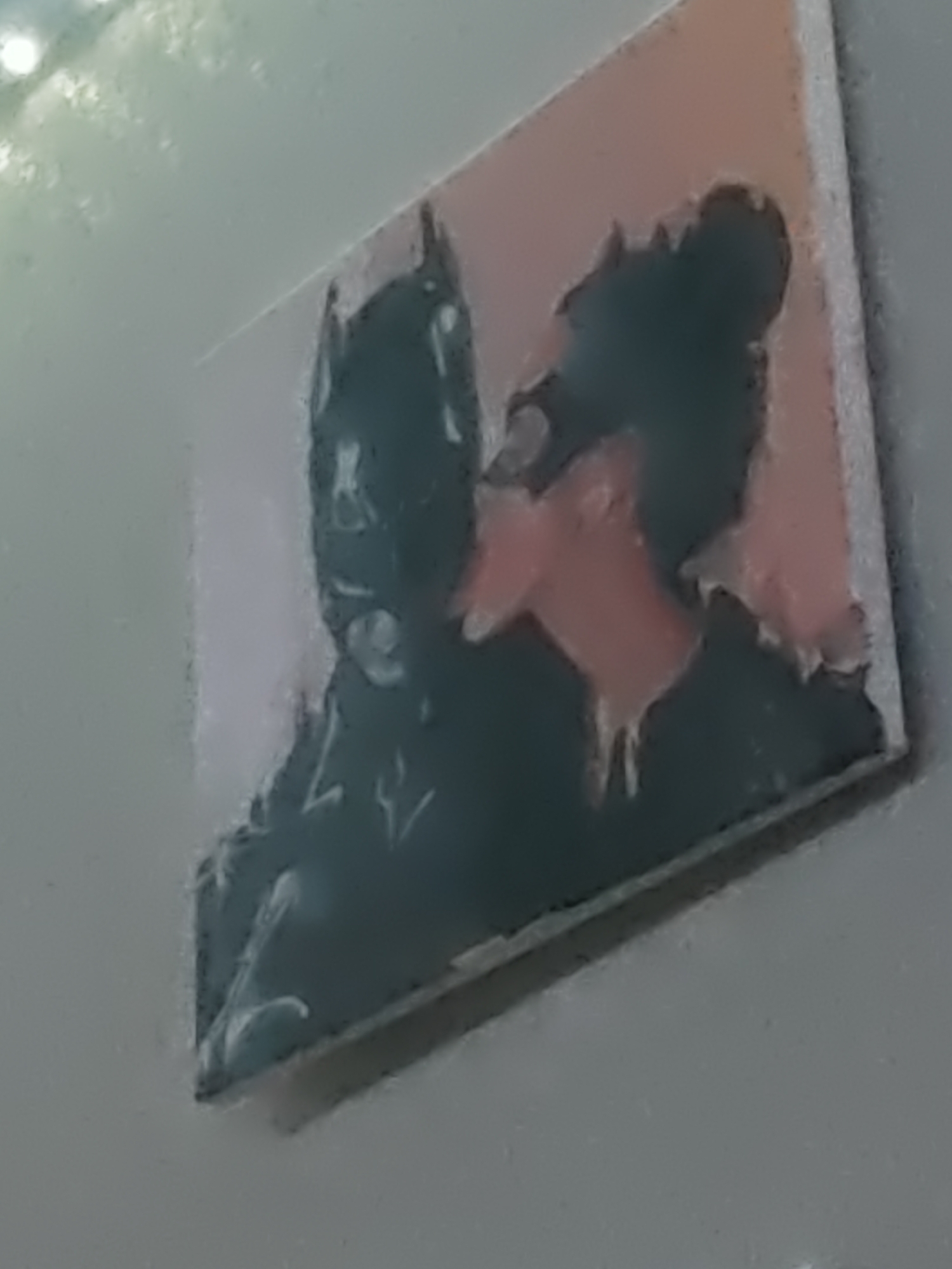 The image is a close-up, slightly blurry photograph of a small, rectangular artwork depicting two figures in silhouette. The figure on the left appears to be Batman, with his distinct cowl and cape visible. The figure on the right is likely Catwoman, suggested by her pointed ears and dark hair styled in a bun. The two figures are positioned in a way that implies an embrace or a close interaction, with their faces almost touching. The artwork has a pinkish-orange background. The overall impression is that this is a piece of merchandise or fan art. The image does not provide any information about its location in the city of Kurganinsk, Russia.