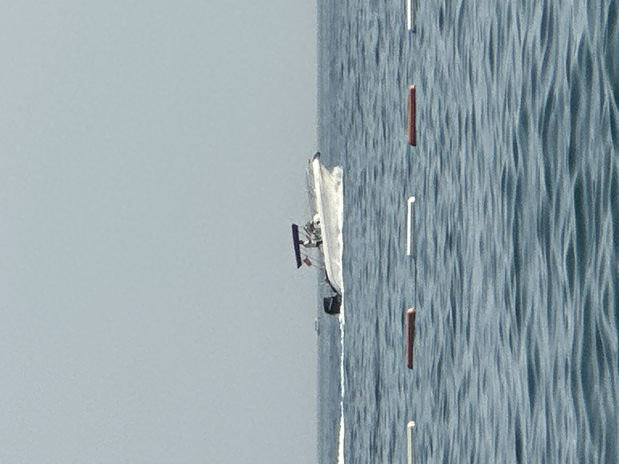 A white motorboat with a blue canopy is moving at speed through the water. A person in a white shirt and dark shorts is visible standing on the boat. The boat is leaving a wake behind it. In the foreground, there are three buoys floating on the surface of the water. The buoys are red and white. The sea is calm and the sky is overcast. The location is the coast of Boreti, Montenegro.