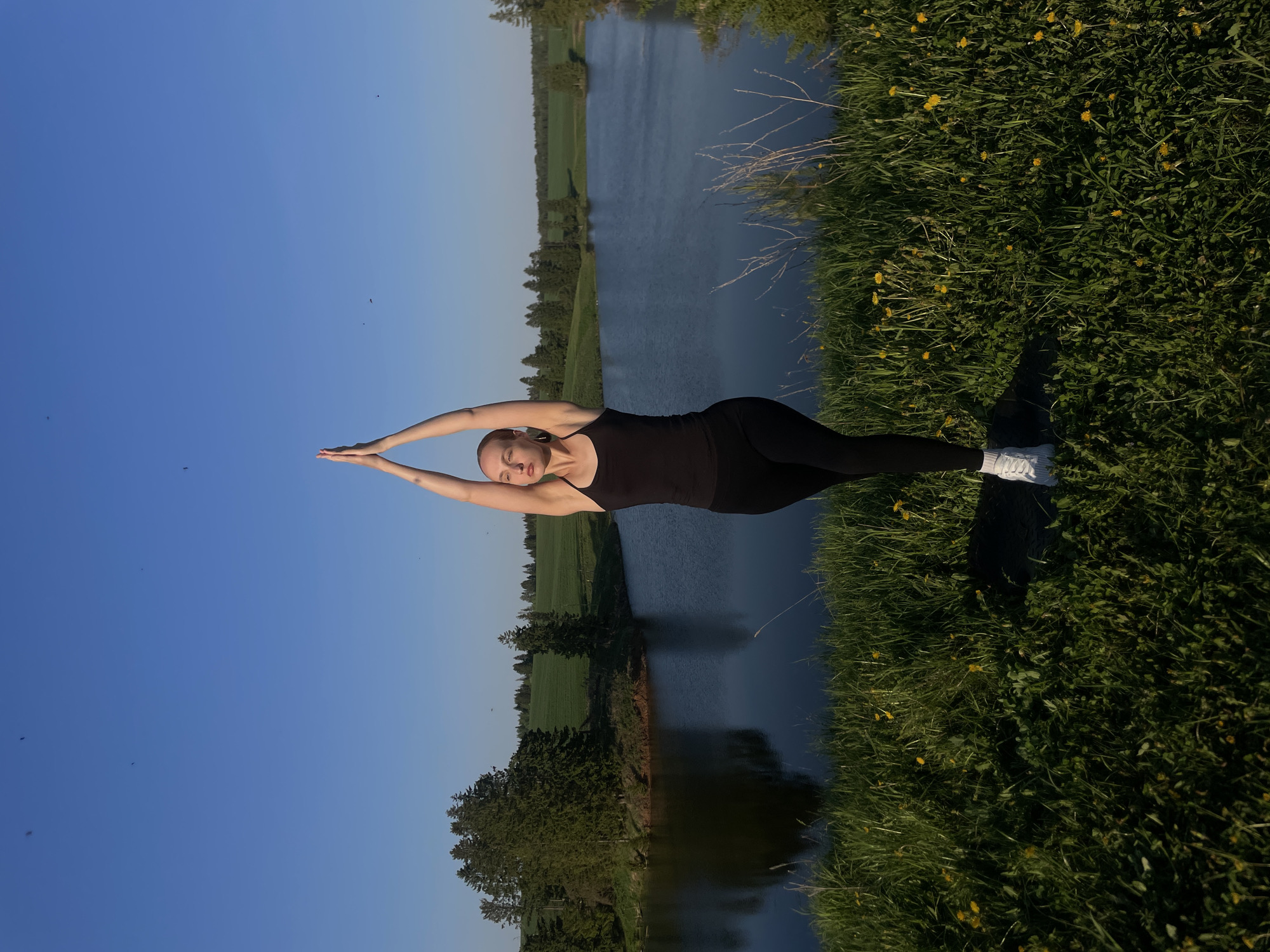 A woman is doing yoga by a body of water. The woman has fair skin, blonde hair, and is wearing a black tank top and black leggings. She is standing on grass and has her arms raised above her head. The body of water is a lake. There are trees and fields in the background. The sky is blue. The location is near Подшивалово, Russia.