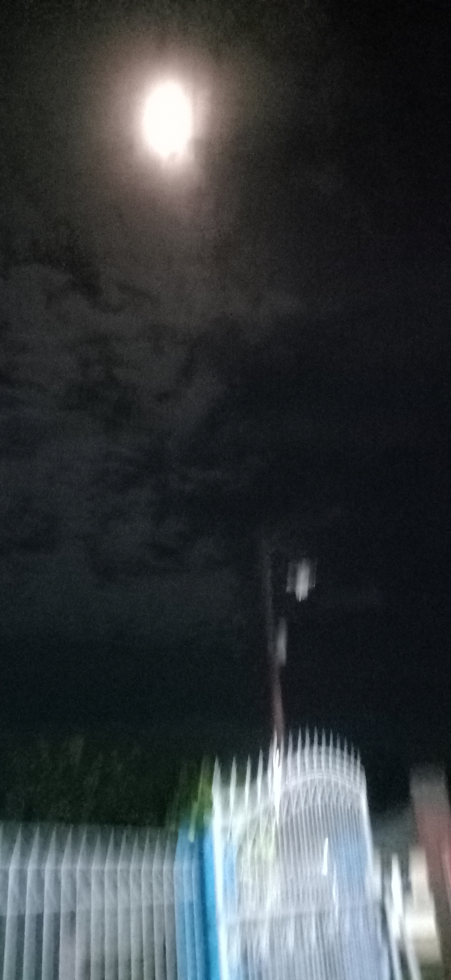 The image shows a night scene with a bright moon in a cloudy sky. In the foreground, a white, ornate fence is visible, with some greenery behind it. A pole with what appears to be a light fixture is also present. The overall impression is of an outdoor urban or suburban setting at night, likely in Lhokseumawe, Indonesia, given the context. The image is somewhat blurry, suggesting it was taken in low light conditions or with movement. There are no people or specific actions depicted.