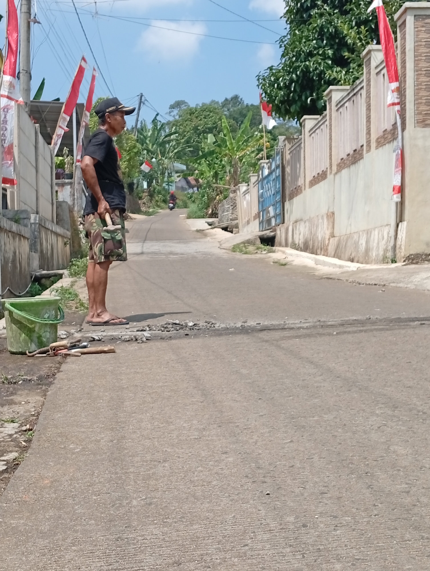 In Cisitu, Indonesia, a man stands on the side of a paved road, holding a tool. He is wearing a black t-shirt, camouflage shorts, a black baseball cap, and flip-flops. In front of him, on the road, are scattered stones and several tools, including a hammer, a trowel, and a shovel. A green bucket sits on the concrete path to his left. The road winds uphill, with houses and lush green trees lining the sides. Red and white Indonesian flags are prominently displayed on poles and on a wall in the background, suggesting a celebration or national holiday. A motorcycle is visible in the distance, heading up the road. The sky is bright blue with scattered white clouds.