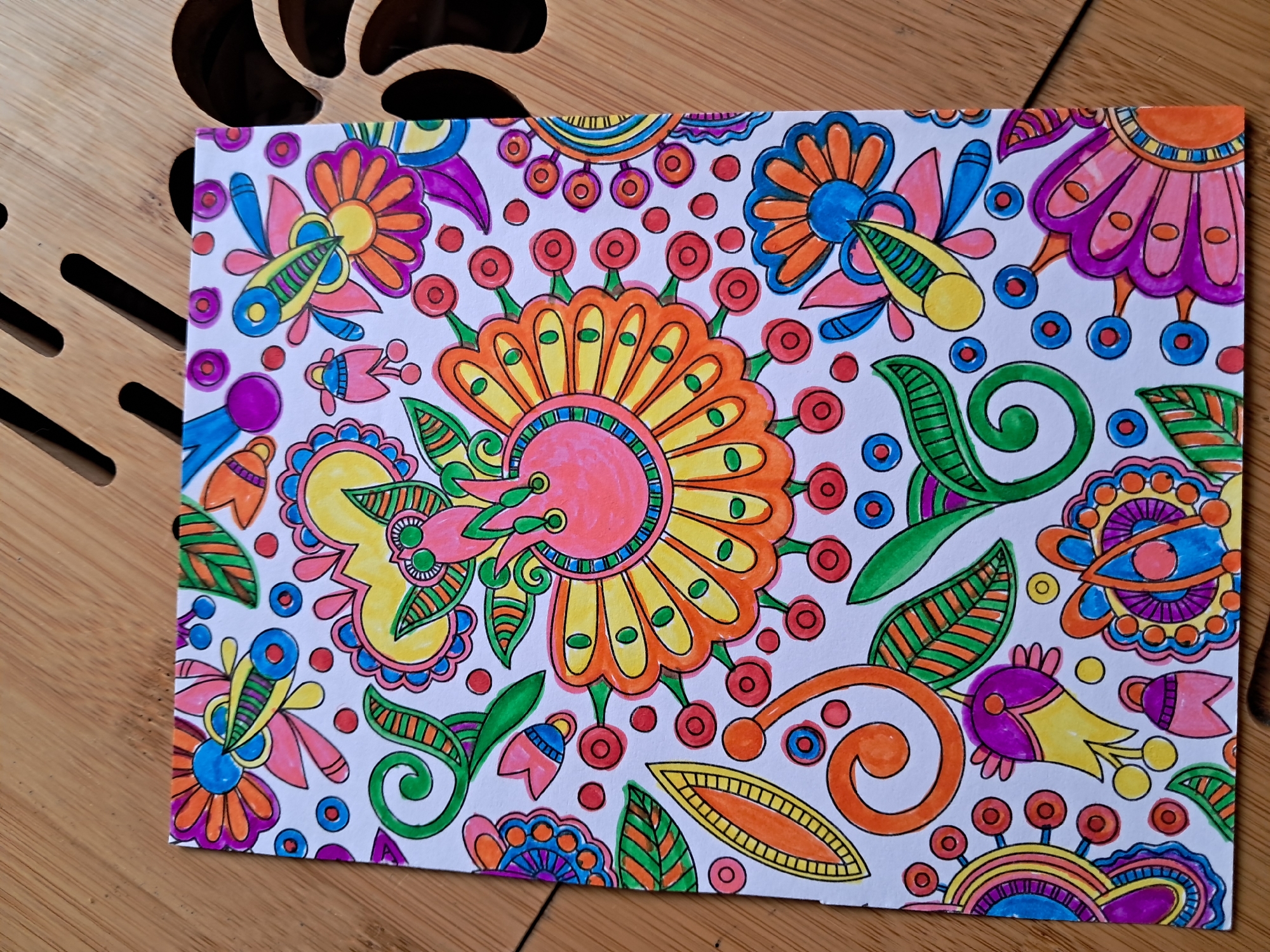 The image displays a piece of paper with a vibrantly colored, abstract floral pattern drawn on it. The drawing features a central, large, sun-like flower with petals in shades of orange and yellow, outlined in black. This central flower is surrounded by various smaller, stylized flowers, leaves, and circular motifs, all rendered in a playful, whimsical style. The colors used are bold and saturated, including pink, purple, blue, green, yellow, and orange. The drawing is executed with clean black outlines.
The paper itself is white and appears to be placed on a wooden surface, possibly a table or a desk. The wooden surface has a slatted design and a light brown color, indicating it is made of wood. Some shadows are cast on the wooden surface from the edges of the paper, suggesting the presence of a light source from above and to the side. The overall impression is of a creative artwork, possibly a coloring page or a doodle, that has been carefully colored in. There is no text visible in the image. The scene is static and focuses solely on the artwork and its immediate surroundings.