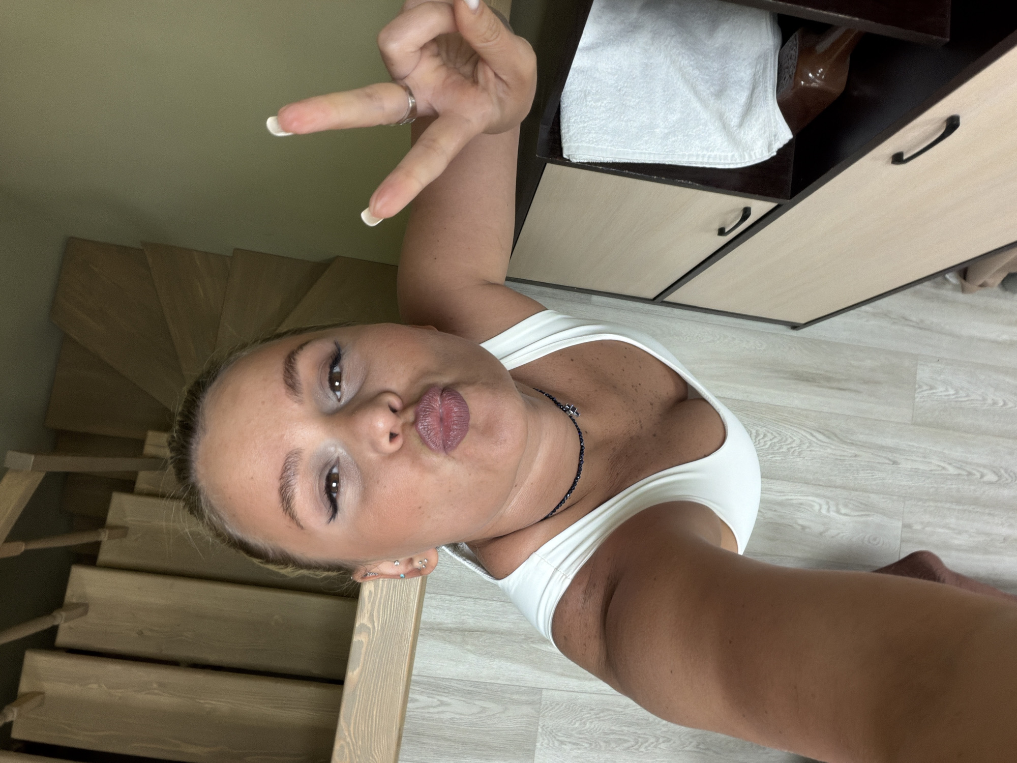 An indoor, vertical selfie captures a young woman posing playfully. She appears fair-skinned with light hair pulled back, dark eyes, and pronounced eyebrows. She is wearing a simple white tank top and a dark beaded necklace. Her right hand is raised, making a peace sign, and her nails are painted with white tips. Her lips are pursed in a "kiss" expression, and multiple earrings are visible on her left ear.
The background on the left features a light-colored wooden staircase with visible treads and railings, indicating an upward ascent. To the right of the woman, a multi-compartment wooden storage unit or cabinet is visible. One open compartment holds a folded white towel and a brown-tinted bottle. Below, several drawers with dark handles are integrated into the unit. The floor is covered with light-colored wooden floorboards. The overall setting suggests a domestic interior, possibly a home or apartment in Sarapul, Russia. The woman's pose conveys a confident and lighthearted mood. No visible text is present in the image.