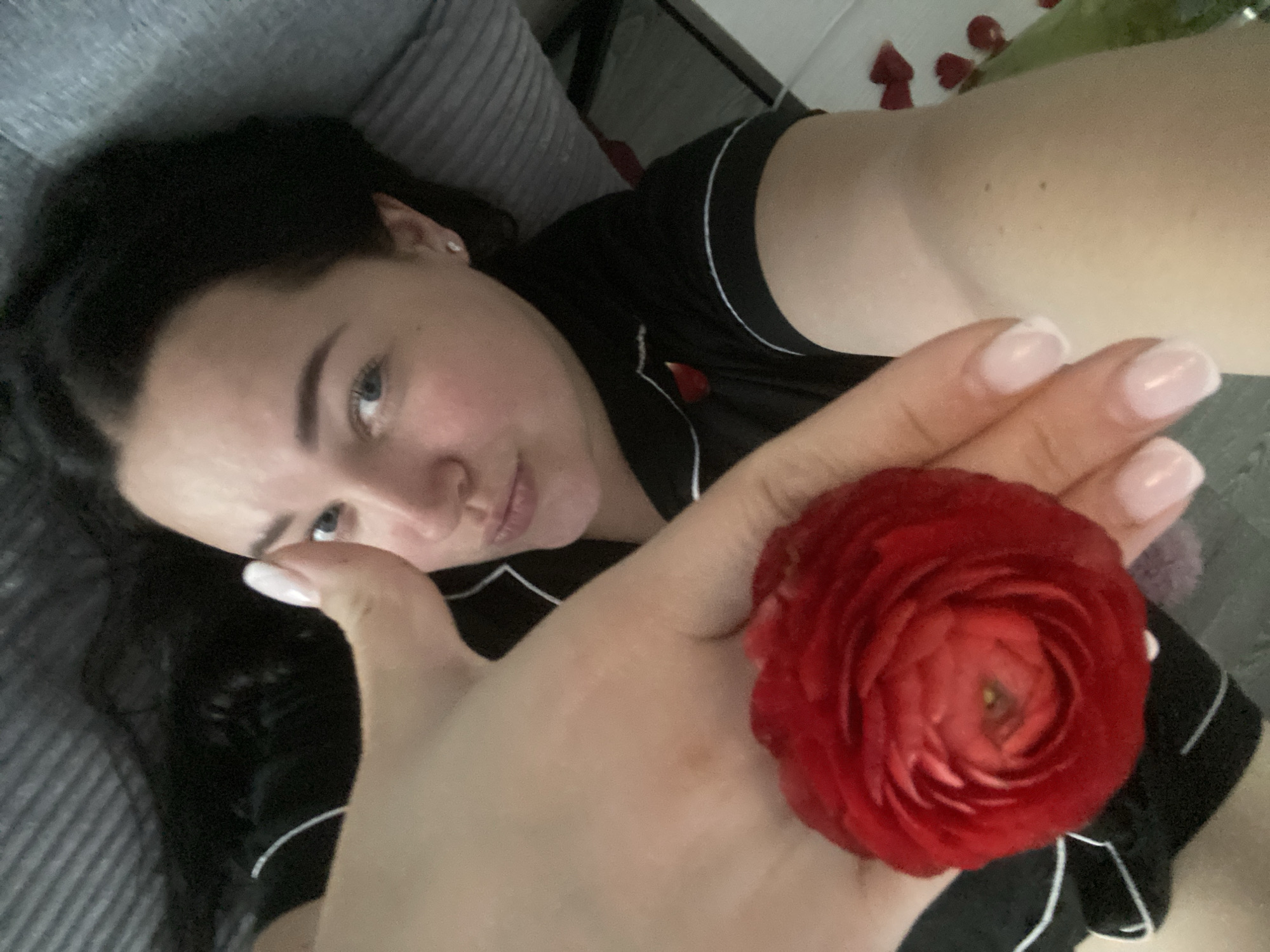 A woman with dark hair and light-colored eyes is captured in a close-up, angled selfie. She is lying down, possibly in a bed, wearing black sleepwear with white piping. Her expression is calm and direct towards the camera. In the foreground, her hand is extended towards the viewer, holding a vibrant red flower, which appears to be a ranunculus, with numerous petals. Her fingernails have a classic French manicure with light pink or nude bases and white tips. Behind her head, a grey striped pillow or cushion is visible. In the background, beyond her outstretched arm, several scattered red rose petals are present, alongside a hint of a green object, possibly a vase or foliage, suggesting a decorative and possibly romantic setting. The scene takes place indoors in Sochi, Russia, conveying a private and relaxed atmosphere. There is no visible text.