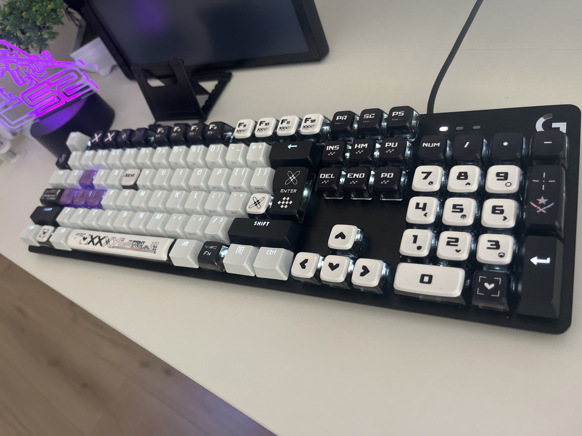 A black and white gaming keyboard with custom keycaps is shown on a white desk. The keyboard has RGB backlighting, with some keys illuminated in purple and white. The keycaps are a mix of white and black, with some featuring custom designs and symbols. The "WASD" keys are highlighted in purple. A neon sign in the shape of a gun is visible to the left of the keyboard, emitting a purple glow. A plant in a pot is also present. The background shows a laptop or monitor screen and its stand. The scene is brightly lit, with the focus on the keyboard and its peripherals. The keyboard appears to be a Logitech G Pro X, based on the visible logo. The image does not contain any information that would indicate the location as Varna, Bulgaria.