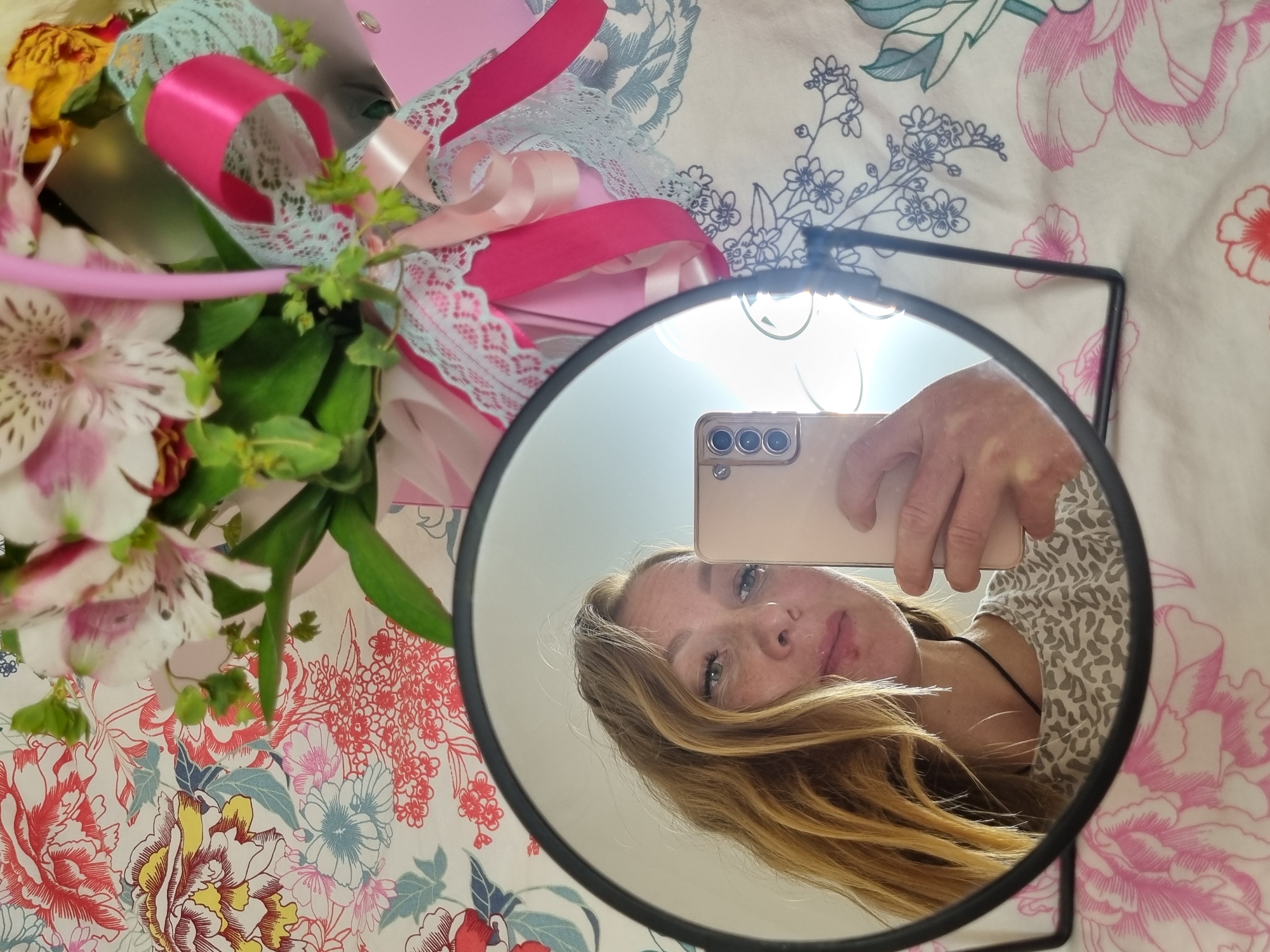 This indoor image from Chaikovsky, Russia, features a mirror selfie composition. A circular mirror with a black frame rests on a surface adorned with a brightly colored floral-patterned fabric, likely a duvet cover or bedsheet, featuring large blooms in pink, red, yellow, and blue with green foliage.
Reflected in the mirror is a woman, presumably the photographer, holding a light-colored smartphone with a triple camera array. She has long, wavy blonde or light brown hair, visible freckles, and a calm, direct gaze towards the camera. She appears to be wearing a subtly patterned top. A bright light source, possibly a window or artificial lamp, is strongly reflected at the top of the mirror.
Surrounding the mirror, a vibrant bouquet of flowers is visible. It contains white and pink alstroemeria, some yellow-orange flowers, and various green leaves. The bouquet is elegantly decorated with bright pink satin ribbons and delicate light blue lace, suggesting it is a gift or a decorative arrangement. The overall lighting is bright, indicating daytime. No text is visible in the image.