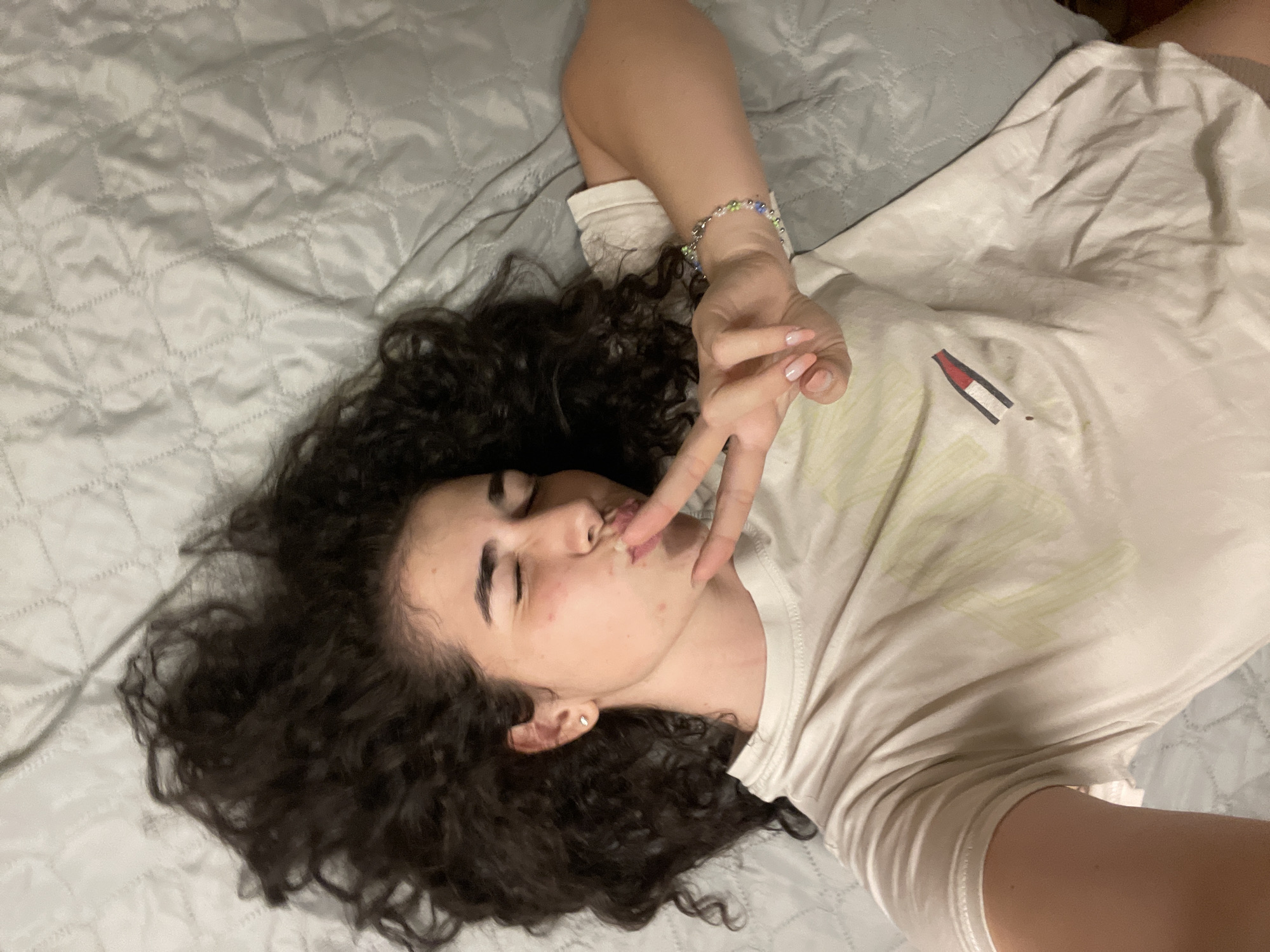 In Sofia, Bulgaria, a young woman with curly dark hair lies on a quilted grey surface, appearing to be a bed or sofa. She is wearing a light-colored t-shirt with a graphic design on the front and a colorful beaded bracelet on her wrist. Her eyes are closed, and she is puckering her lips while holding up two fingers in a peace sign. The lighting suggests an indoor setting, possibly a bedroom.