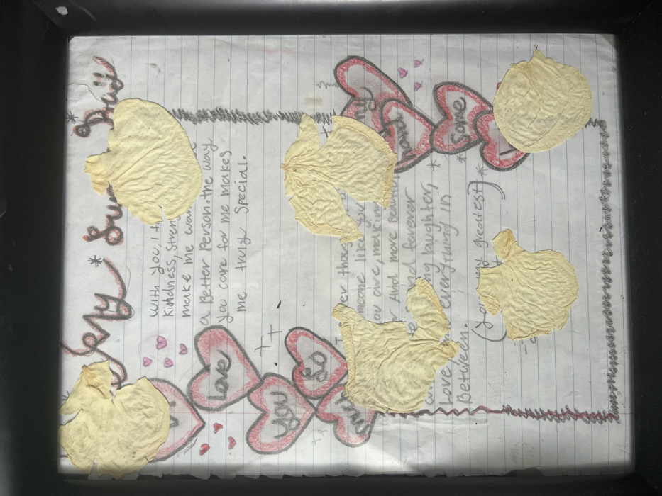A handmade piece of art features a handwritten love letter on lined paper. The text expresses gratitude and affection, referencing kindness, strength, and a desire to be a better person. Embellishments include drawn red hearts with messages like "Love You So Much" and decorative borders along the top and bottom. Dried, pale yellow flower petals are scattered across the page, adding a natural and textural element. The entire piece is encased within a black frame.
