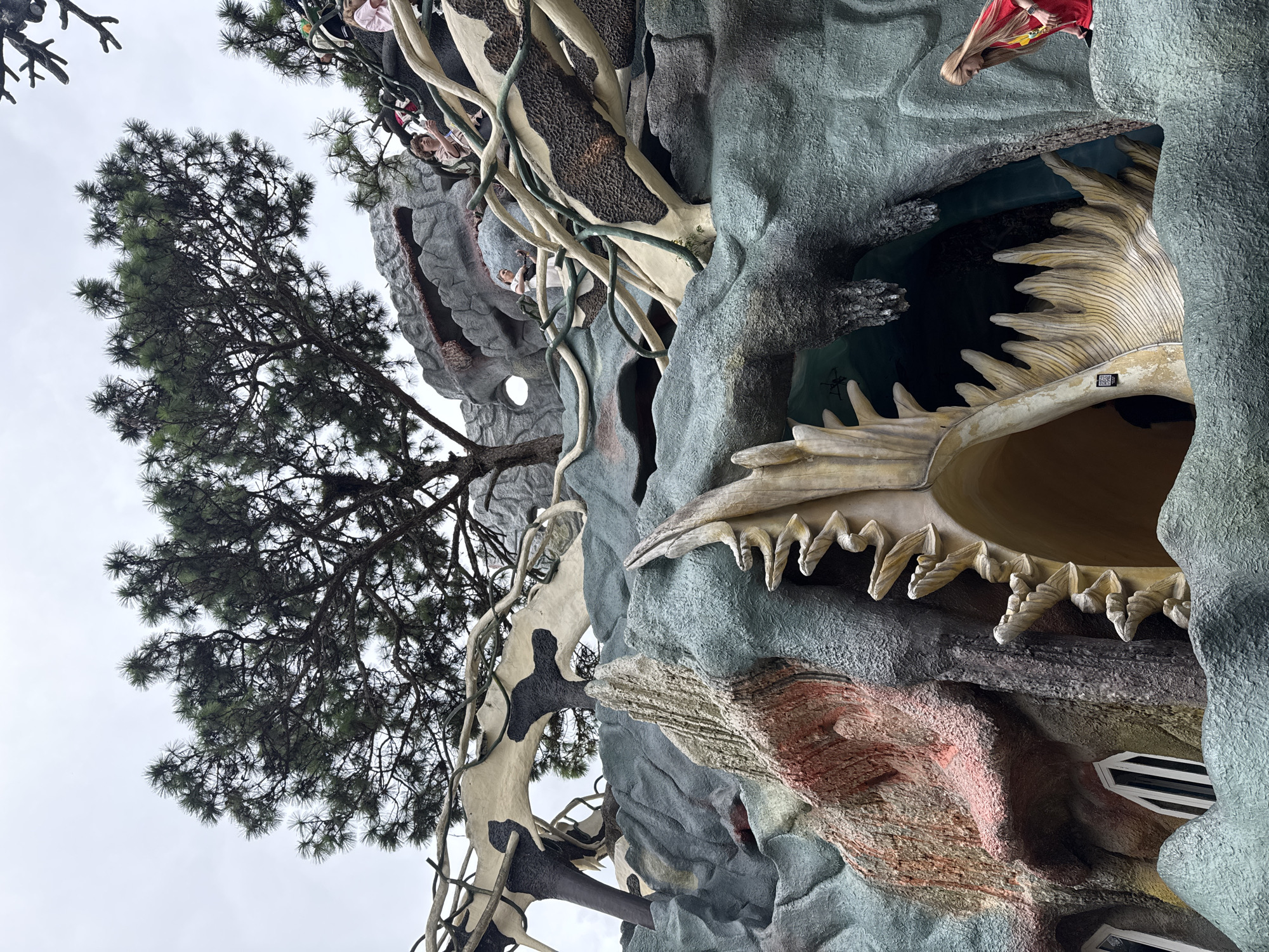 This vertically oriented image captures a fantastical, organic architectural structure, likely the "Crazy House" in Da Lat, Vietnam, as seen from a low angle looking upwards. The building features an unconventional design with irregular, rock-like textures in muted greens, grays, and reddish-browns, mimicking natural formations and integrated with faux tree trunks and winding root-like elements.
Several people are visible interacting with the structure. In the upper left, a person in a pink top can be seen on a balcony. Another individual in a white top is standing on a branch-like protrusion further down the building. Most prominently, a person with long blonde hair and a red top is peering out from an opening, their reflection subtly visible on the surface below.
A large, stylized opening on the right side resembles a giant, skeletal animal's mouth or a massive seashell, complete with sharp, irregular teeth-like protrusions. A small, dark entrance is visible behind these teeth. Windows of various shapes, including a multi-paned rectangular one, are set into the textured walls.
A prominent pine tree with dense foliage occupies the left side of the frame, its dark silhouette contrasting against the overcast, light gray sky. The cloudy weather suggests a daytime shot, but without direct sunlight. A small QR code is visible on one of the teeth-like structures. The scene conveys a sense of wonder and surrealism, characteristic of the unique architecture of the Crazy House.