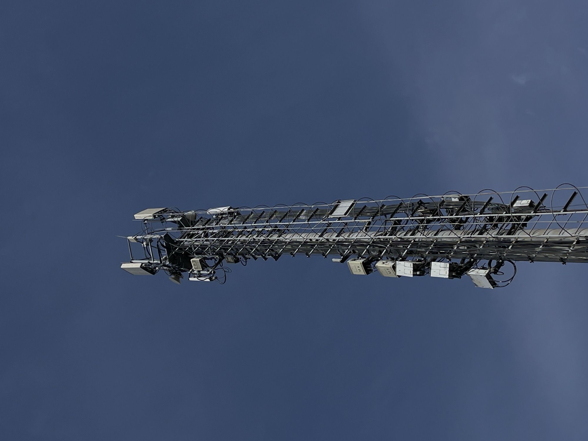 A low-angle, horizontal shot depicts a section of a cellular communication tower against a clear, dark blue sky. The tower section is positioned along the horizontal axis of the frame. It is constructed of metal with a lattice-like pattern and features numerous small, rectangular, gray boxes attached along its length. These boxes are connected to the tower by black cables. The tower structure is detailed, revealing the complexity of the communication equipment. The background consists only of the uniform, featureless dark blue sky.