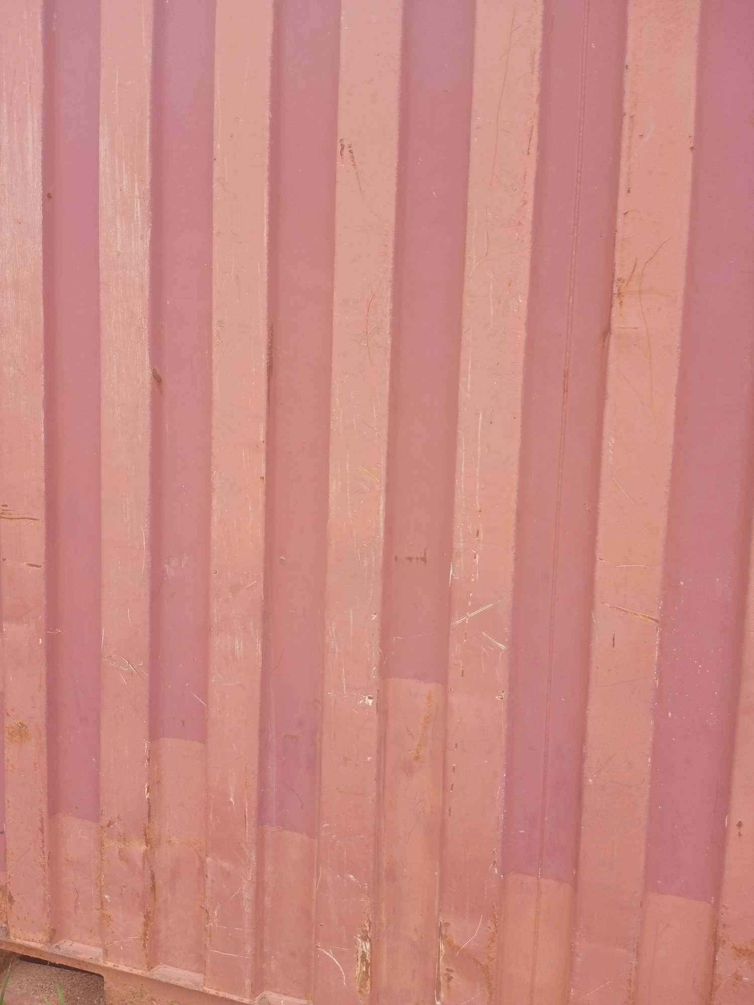 The image shows a close-up view of the corrugated exterior of a weathered red shipping container. The surface exhibits a textured pattern of vertical ridges and valleys, with visible signs of wear such as scratches, scuffs, and some minor rust spots. The lighting suggests an outdoor environment, casting subtle shadows that accentuate the corrugated texture. The overall impression is one of a utilitarian and durable industrial material. There are no people, animals, or specific landmarks visible in the frame, making it impossible to ascertain the exact location within Jalingo, Nigeria, or any specific activity.