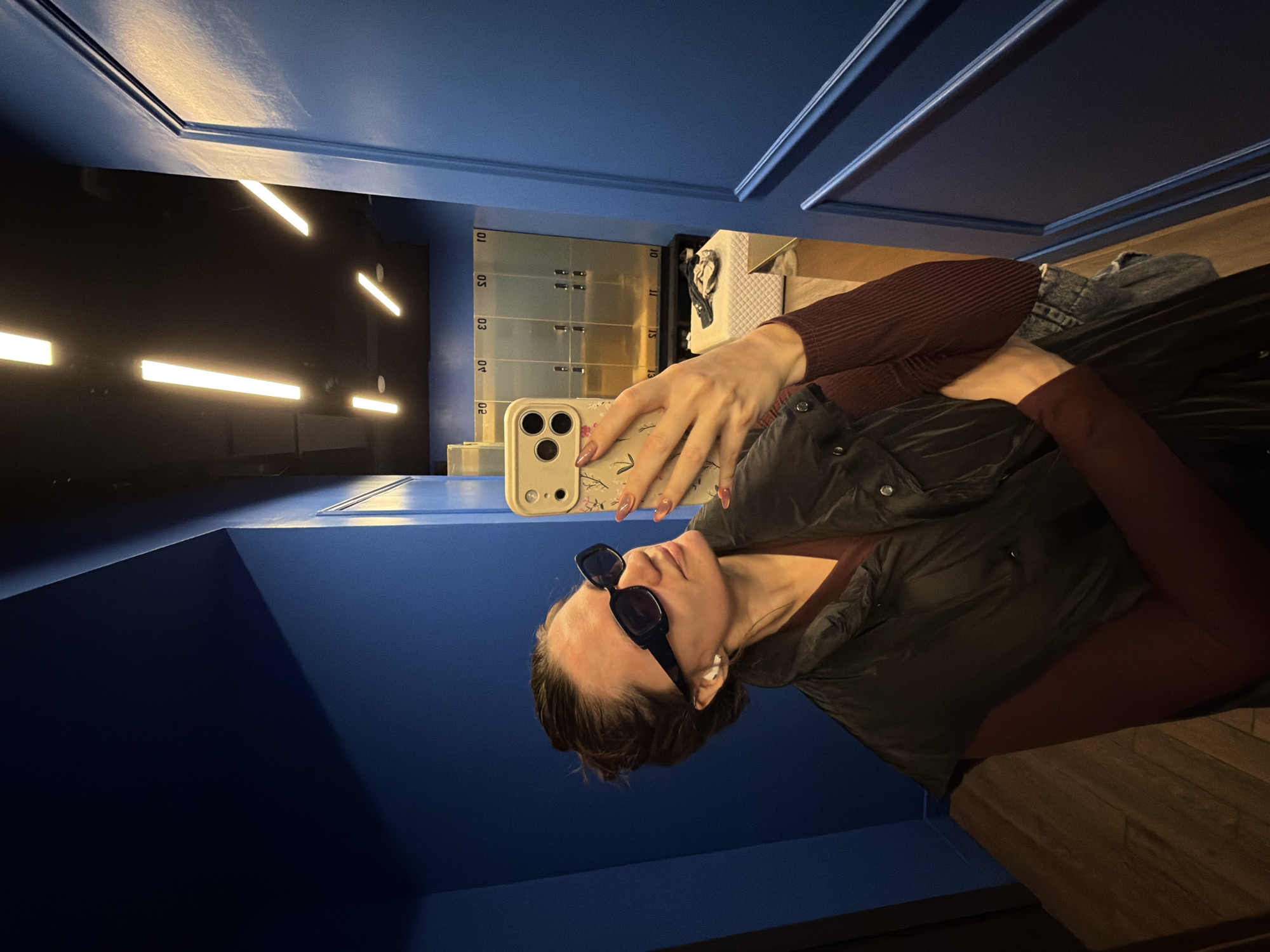 The image displays an indoor mirror selfie taken by a person in what appears to be a modern facility in Belgrade, Serbia. The subject is a female-presenting individual, visible from the chest up, posing with a smartphone held up to capture their reflection. They are wearing dark, rectangular sunglasses and a dark puffy vest or jacket over a reddish-brown long-sleeved top. A small white stud earring is visible in their right ear, and their fingernails are painted a light, neutral color.
The setting features prominent deep blue walls, some of which have decorative moldings. The ceiling or upper wall area is dark, with several bright, linear LED light fixtures arranged in an abstract pattern. In the background, to the left of the subject, there is a bank of silver-colored lockers with visible numbers such as "01," "02," "03," "05," "10," "11," "13," and "15." To the right of the lockers, a white, quilted surface, possibly part of a bench or seating, and some folded denim clothing are visible. The floor is constructed from light brown wooden planks. The overall impression is that of a contemporary locker room, gym, or a public area designed with modern aesthetics and lighting. The time of day is not discernible due to the artificial lighting.