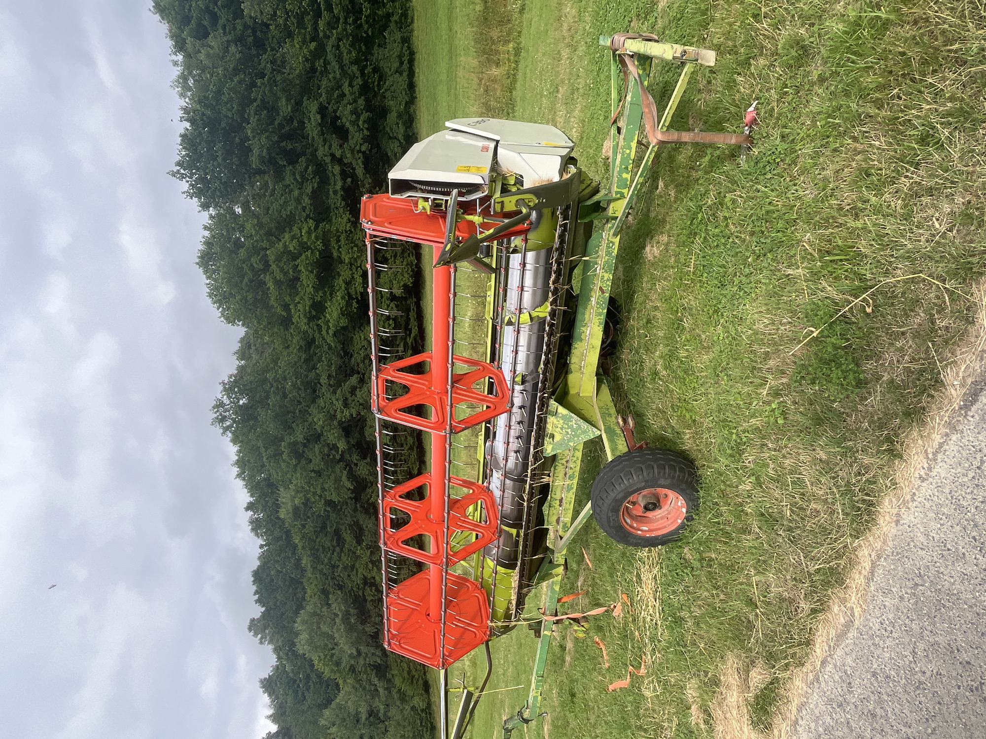 The image shows a farm implement, likely a combine harvester header, parked on a grassy field in Wiera, Germany. The implement is primarily green and orange, with a large reel at the front and a conveyor system behind it. A single wheel with a red rim is visible on the right side of the implement. The background consists of a dense line of trees and a cloudy sky. The scene appears to be outdoors, with no people visible. The grass suggests it is a rural or agricultural setting. The lighting indicates it could be daytime, but the cloudy sky suggests overcast weather. No specific activity or event is taking place; the implement appears to be stationary. The visible text on the implement is "CLAAS C390".