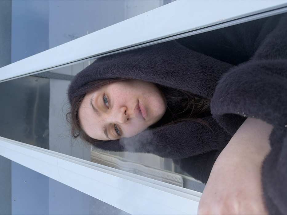 An adult female is depicted in a medium shot, positioned within or leaning out of an open window frame in Chaikovsky, Russia. She has brown hair, light-colored eyes, and a fair complexion. She wears a dark, possibly charcoal-colored, hooded faux fur garment and a dark turtleneck beneath. A plume of white vapor or smoke is visible emanating from her mouth, indicating an exhalation. Her lips are slightly parted, and her gaze is directed towards the viewer. The lower left portion of the image shows a bare segment of her leg, likely a thigh, above the dark garment. The white window frame diagonally bisects the upper part of the image, with clear glass panes revealing a light blue/grey sky and indistinct background elements, consistent with an outdoor environment.