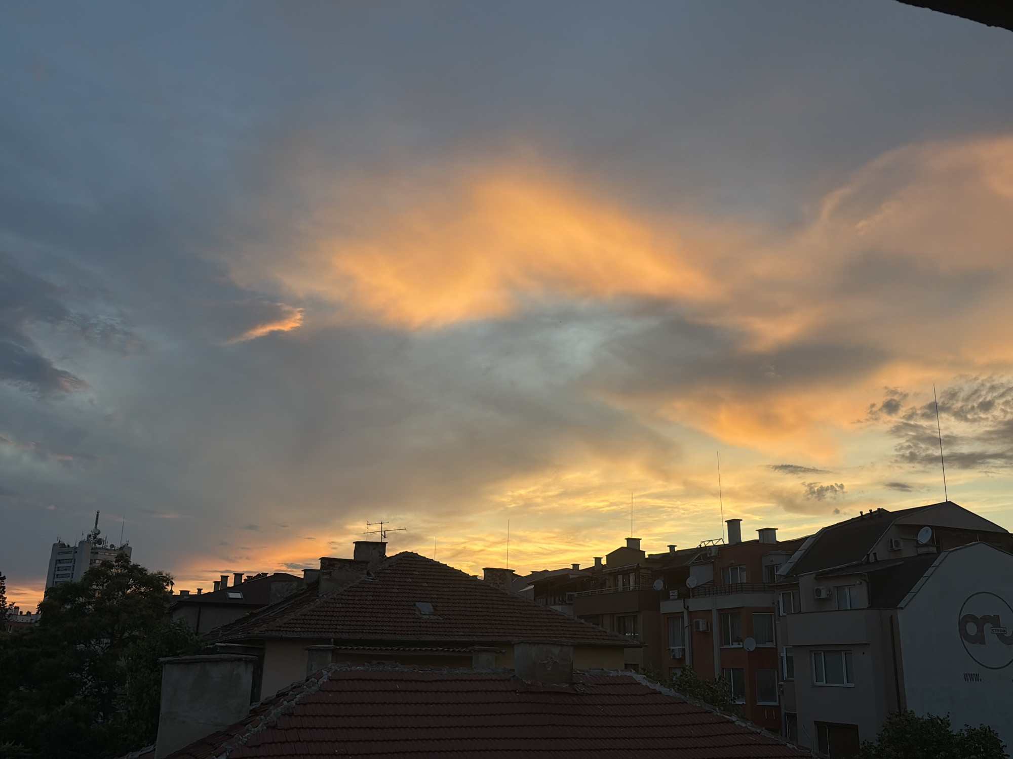 The image captures a cityscape at dusk, dominated by a dramatic sky filled with clouds. The clouds are illuminated with warm hues of orange and yellow, indicating the setting sun, while other parts of the sky remain a cooler, muted blue-gray. The foreground is filled with the rooftops of buildings, featuring red tile roofs, chimneys, and satellite dishes. In the background, a taller, more modern building with an antenna array stands out against the colorful sky. The overall scene depicts an urban environment as day transitions into night, with a focus on the interplay of light and shadow on the architecture and the sky.