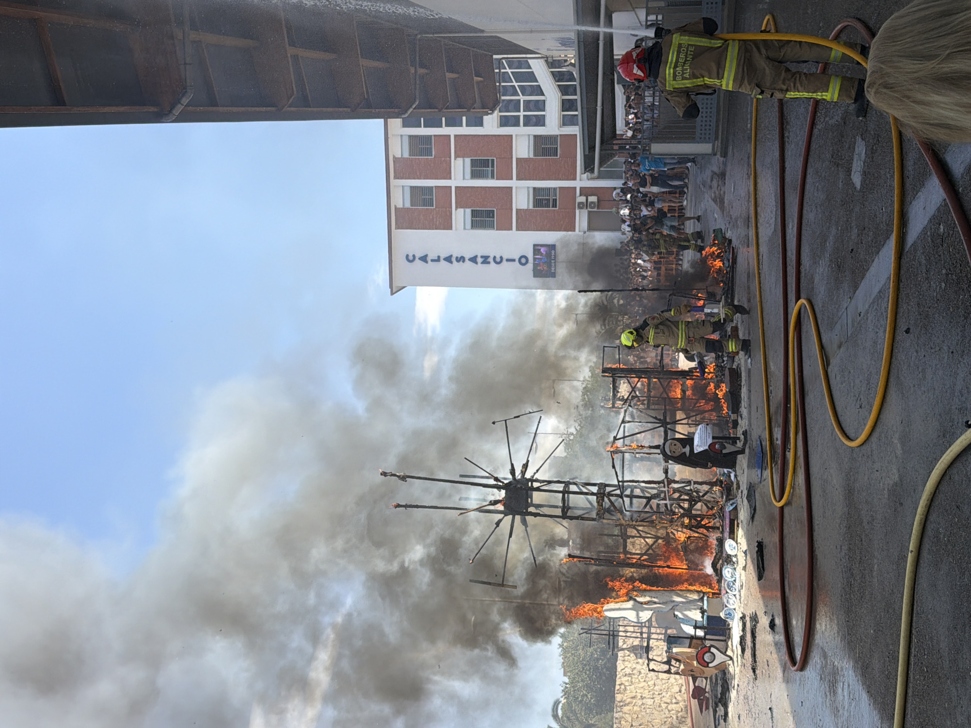 The image depicts a street scene with a large structure engulfed in flames and thick black smoke billowing into a clear blue sky. In the foreground, two firefighters in protective gear are present. One firefighter, positioned on the right, is operating a hose, directing water towards the fire. The other firefighter is visible to the left, standing near the burning structure. Several lengths of fire hose are laid out on the wet asphalt. In the background, a multi-story building with "CALASANCIO" written vertically in large blue letters is visible. A crowd of onlookers stands behind a railing, observing the fire. The environment appears to be an urban setting, possibly in front of an educational institution, given the building's signage. The ground is wet, indicating water has been applied to the fire.