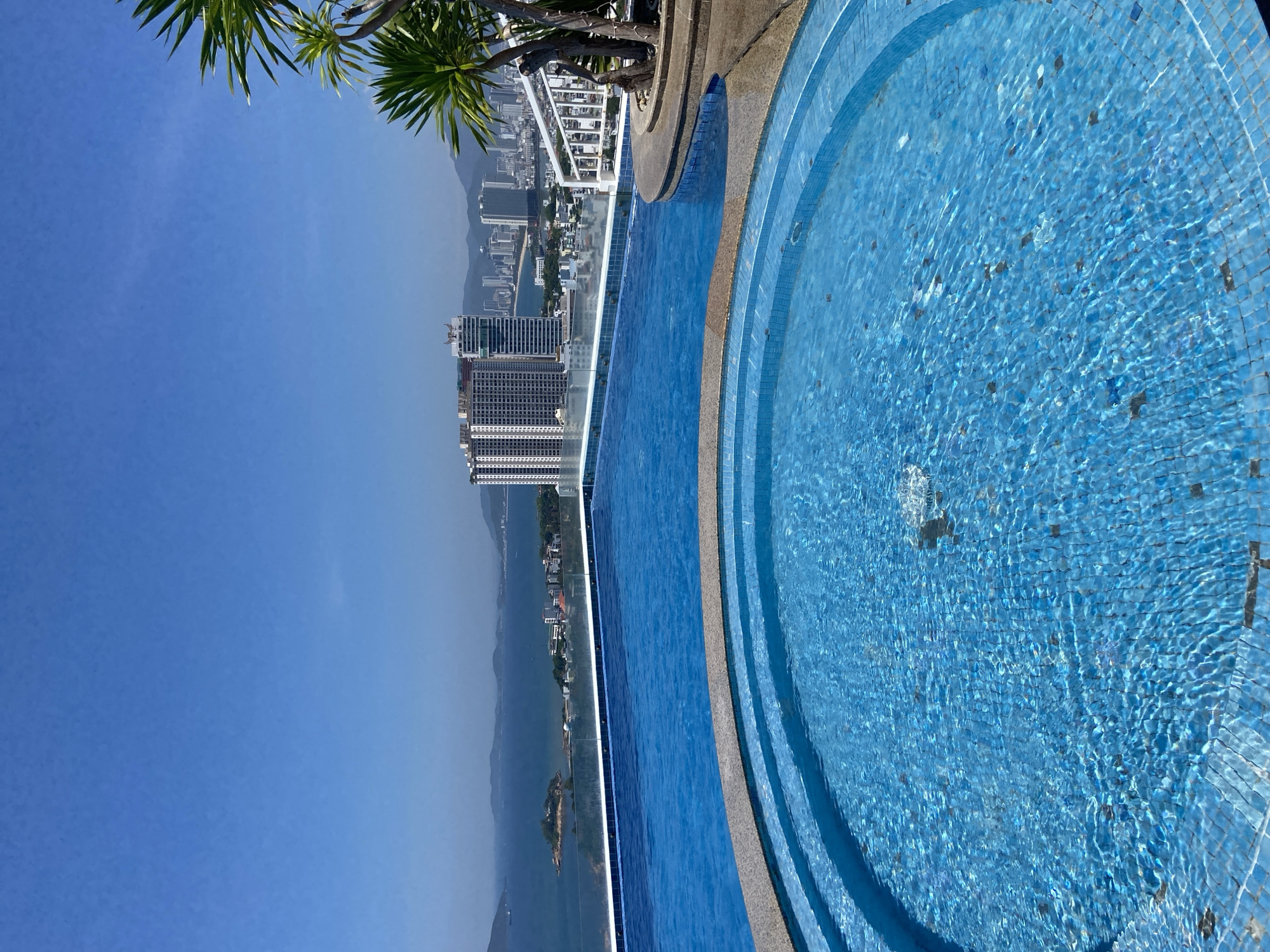 This media file captures a high-angle view of a bright, azure swimming pool, likely an infinity pool given its placement overlooking a cityscape and ocean. The pool is tiled in small, shimmering blue tiles, with distinct concentric steps leading down into the water. Light refracts across the pool's surface, creating a dynamic, rippling texture. A small fountain or water feature is visible, gently disturbing the water's surface in the foreground.
Beyond the pool, a cityscape unfolds, characterized by numerous modern high-rise buildings. The city appears to stretch towards a hazy horizon, where mountains rise in the distance. The sea is visible between the city and the mountains, with a small island just offshore. Lush green foliage, appearing to be palm fronds, overhangs the scene from the top left, suggesting a tropical or sub-tropical setting.
The sky is a clear, vibrant blue, indicating a sunny day. The lighting suggests it is daytime, likely mid-morning or early afternoon. The overall impression is one of a serene and luxurious setting, perhaps a hotel or resort in a coastal city. There is no visible text in the image. The scene evokes a sense of tranquility and leisure. The orientation of the image is sideways.