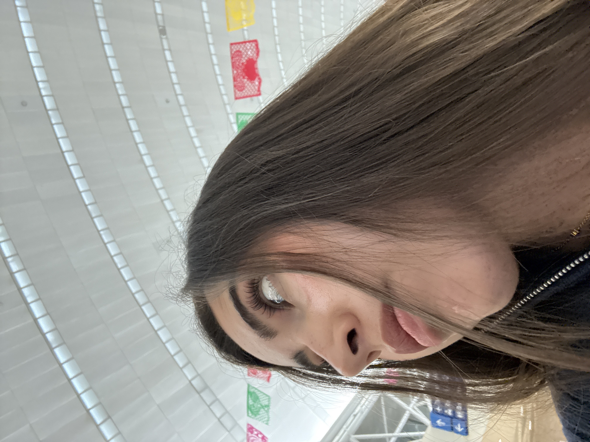 A close-up, tilted shot focuses on a young woman with long, straight, light brown hair, fair skin, and dark eyebrows. Her gaze is directed upwards. A portion of her face is visible including one eye, the nose, and part of the mouth. She wears dark clothing with a visible zipper. The background features a white, grid-patterned ceiling. Below are strings of colorful paper decorations, with green, red, and yellow colors evident. A sign with blue arrows indicating direction is also visible. This is taken at the San Antonio International Airport in San Antonio, Texas.