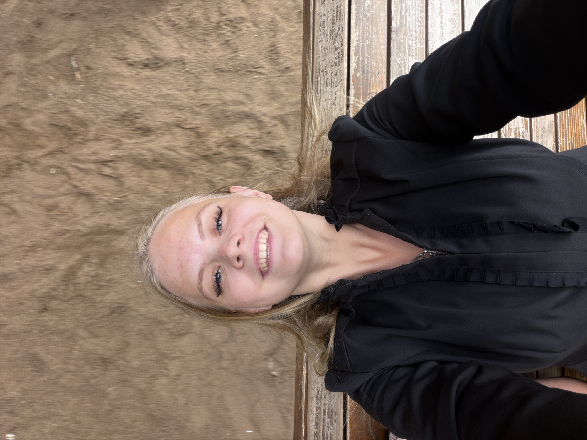 A young woman with blonde hair and freckles is taking a selfie outdoors. She is wearing a black jacket with ruffled trim and a silver necklace. She is sitting on a wooden bench, with a sandy or dirt wall behind her. The image is taken from a slightly low angle, looking up at the woman. The woman is smiling and looking at the camera. The location is not identifiable as Izhevsk, Russia.