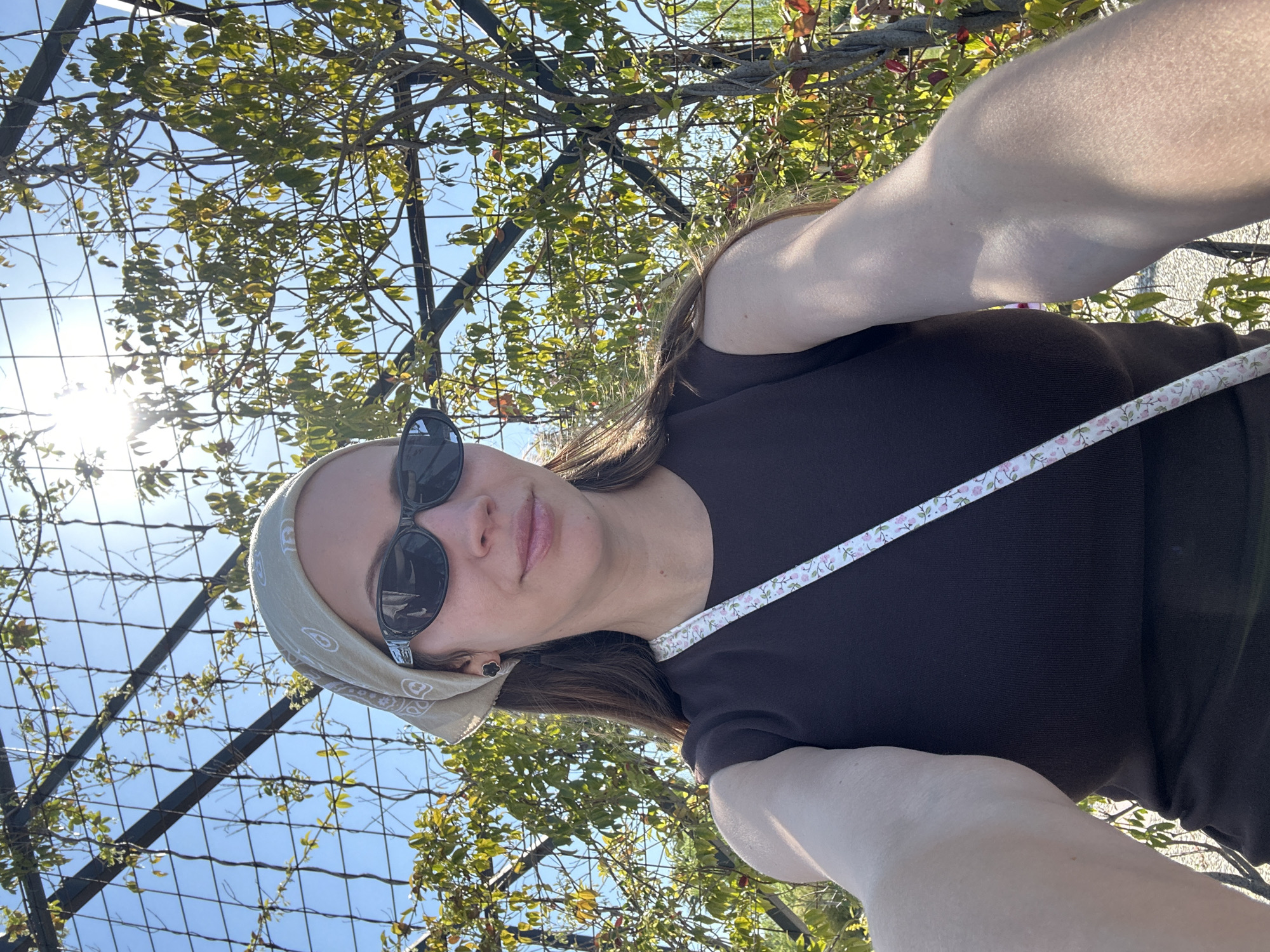 A person takes a vertical selfie outdoors, positioned in the lower right of the frame, looking up towards the camera. She wears dark sunglasses, a light beige bandana with a subtle pattern around her head, and a dark brown, possibly ribbed, short-sleeved top. A white strap with a small pink and green floral pattern crosses diagonally across her chest. Her brown hair is visible beneath the bandana and over her shoulders.
The background features a metal grid structure, likely a pergola or trellis, heavily adorned with lush green climbing plants and some scattered reddish leaves. Patches of clear blue sky are visible through the gaps in the foliage and grid. A strong sun flare emanates from the upper left quadrant of the image, indicating bright, direct sunlight. The lighting is intense and sunny, suggesting midday or early afternoon. The setting appears to be an outdoor garden or park area, consistent with a sunny location like Fethiye, Turkey. No visible text is present.