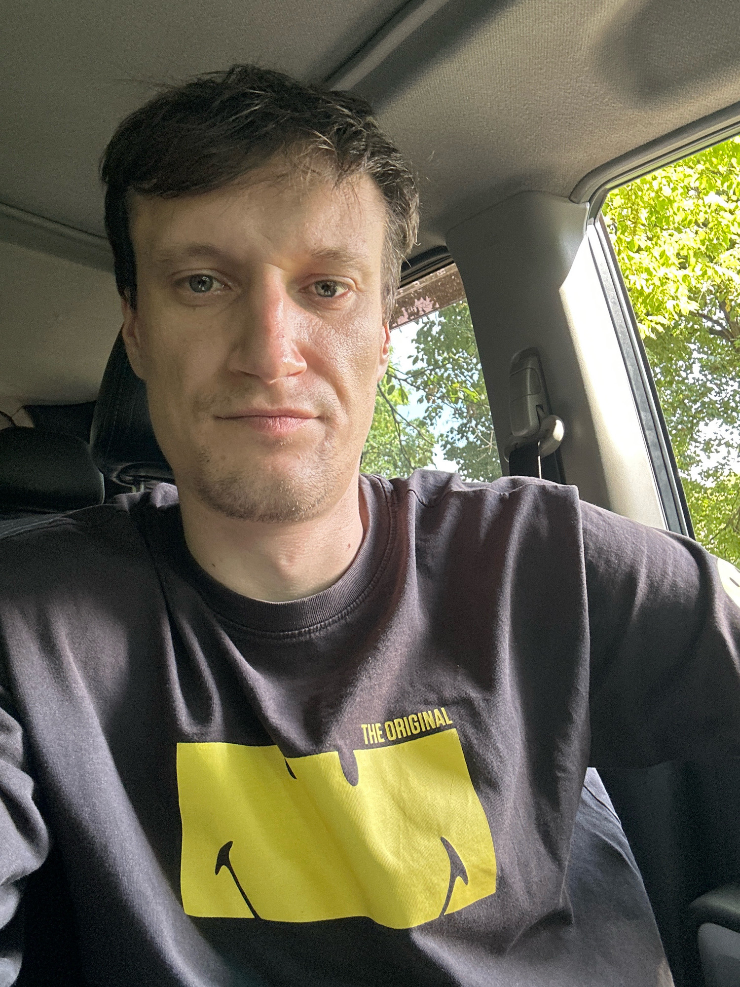 A man is sitting in the driver's seat of a car, looking directly at the camera. He has brown hair and is wearing a black t-shirt with a yellow smiley face and the words "THE ORIGINAL" printed on it. The car's interior is visible, including the headliner and a portion of the seat. Outside the car, through the window, green trees and foliage are visible, suggesting an outdoor, possibly wooded, location. There is no specific information about the location being in Shatava, Ukraine, or any city in Ukraine.