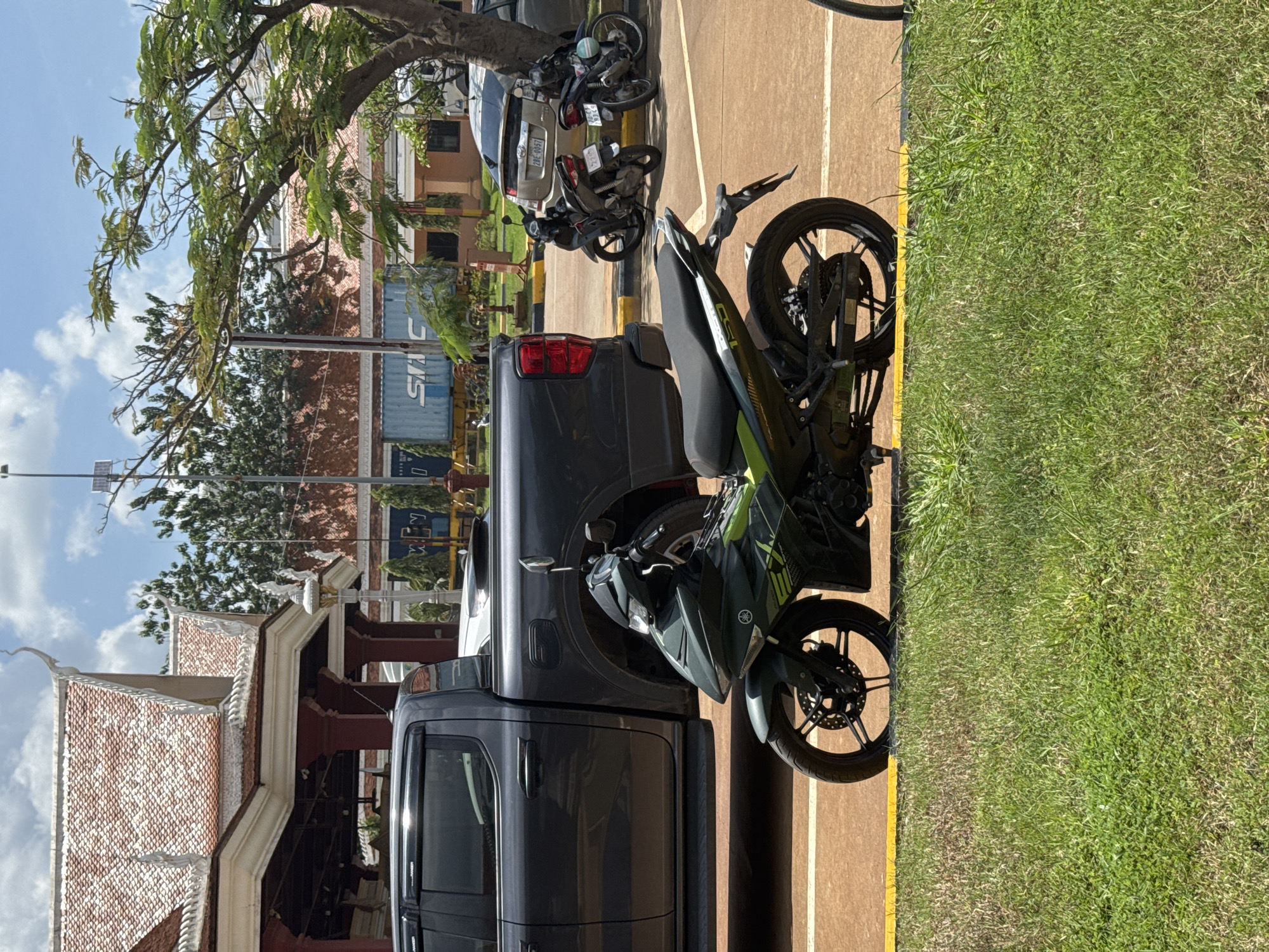 In Bavet Quarter, Cambodia, a dark grey pickup truck is parked next to a green and black motorcycle. The motorcycle is positioned in the foreground, facing left, with its front wheel turned. Behind the pickup truck, two more motorcycles are visible, parked to the right. Further in the background, a silver car is parked near a tree. Buildings with traditional tiled roofs and red accents are visible behind the vehicles. A blue shipping container and other structures indicate an industrial or border crossing area. The scene is set outdoors under a bright, partially cloudy sky, with a grassy area in the immediate foreground.