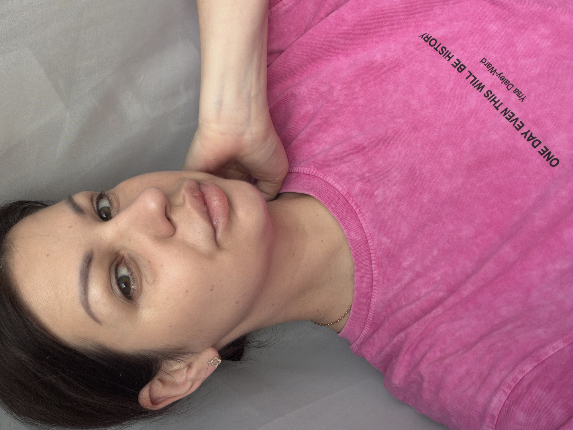 The image shows a close-up, waist-up, selfie-style shot of a woman with dark hair, brown eyes, and light skin. She is wearing a bright pink t-shirt with text printed on it in black. The text reads "ONE DAY EVEN THIS WILL BE HISTORY" followed by "Yrsa Daley-Ward". She is resting her right hand on the back of her neck, and her left hand is not visible. She is wearing a small, diamond-shaped earring in her left ear and a thin gold chain around her neck. The background appears to be a light gray, textured surface, possibly a fabric backdrop. The overall lighting is soft and diffused. The image does not contain any visual cues that indicate its location in Sochi, Russia.