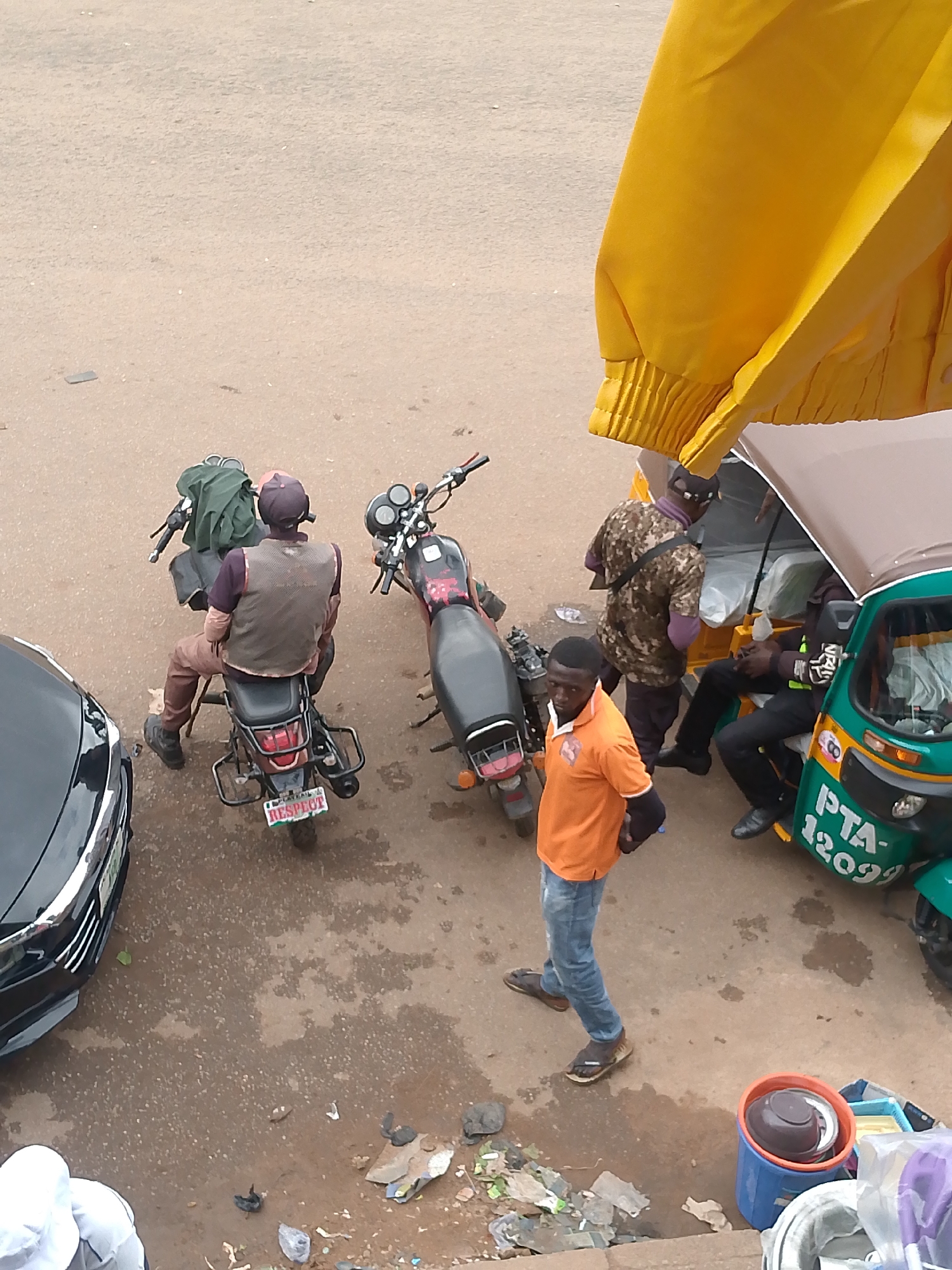 In Bukuru, Nigeria, a scene unfolds on a dusty, light-brown paved surface. The image captures several individuals and their modes of transport.
In the foreground, a man in an orange t-shirt and blue jeans stands facing left, his body slightly turned towards the viewer. He wears brown sandals and appears to be holding a dark object in his right hand.
To his left, a black car is partially visible, its sleek design contrasting with the rugged surroundings.
Behind the man in orange, a motorcycle with a red and black body is parked. A man wearing a brown vest over a purple shirt and brown trousers sits on the motorcycle. A green bag is strapped to the back of the motorcycle. The motorcycle has a license plate that reads "RESPECT".
Further back, another motorcycle, this one with a red and black frame, is parked to the right of the first motorcycle.
To the right of the motorcycles, a green and white three-wheeled auto-rickshaw is parked. Two men are seated inside the rickshaw. One man, wearing a camouflage shirt and a purple undershirt, is looking to the left. The other man, wearing a black t-shirt with reflective stripes and dark trousers, is looking forward. The rickshaw has "PTA-12029" written on its side.
A yellow awning or fabric is visible in the upper right portion of the image, extending over the auto-rickshaw.
The ground is littered with debris, including crumpled paper, plastic bags, and what appear to be discarded food items or waste. There are also two buckets, one blue and one orange, with items inside them, near the bottom right of the frame.
The overall environment suggests a busy street or roadside location, possibly a transportation hub or market area in Bukuru, Nigeria.