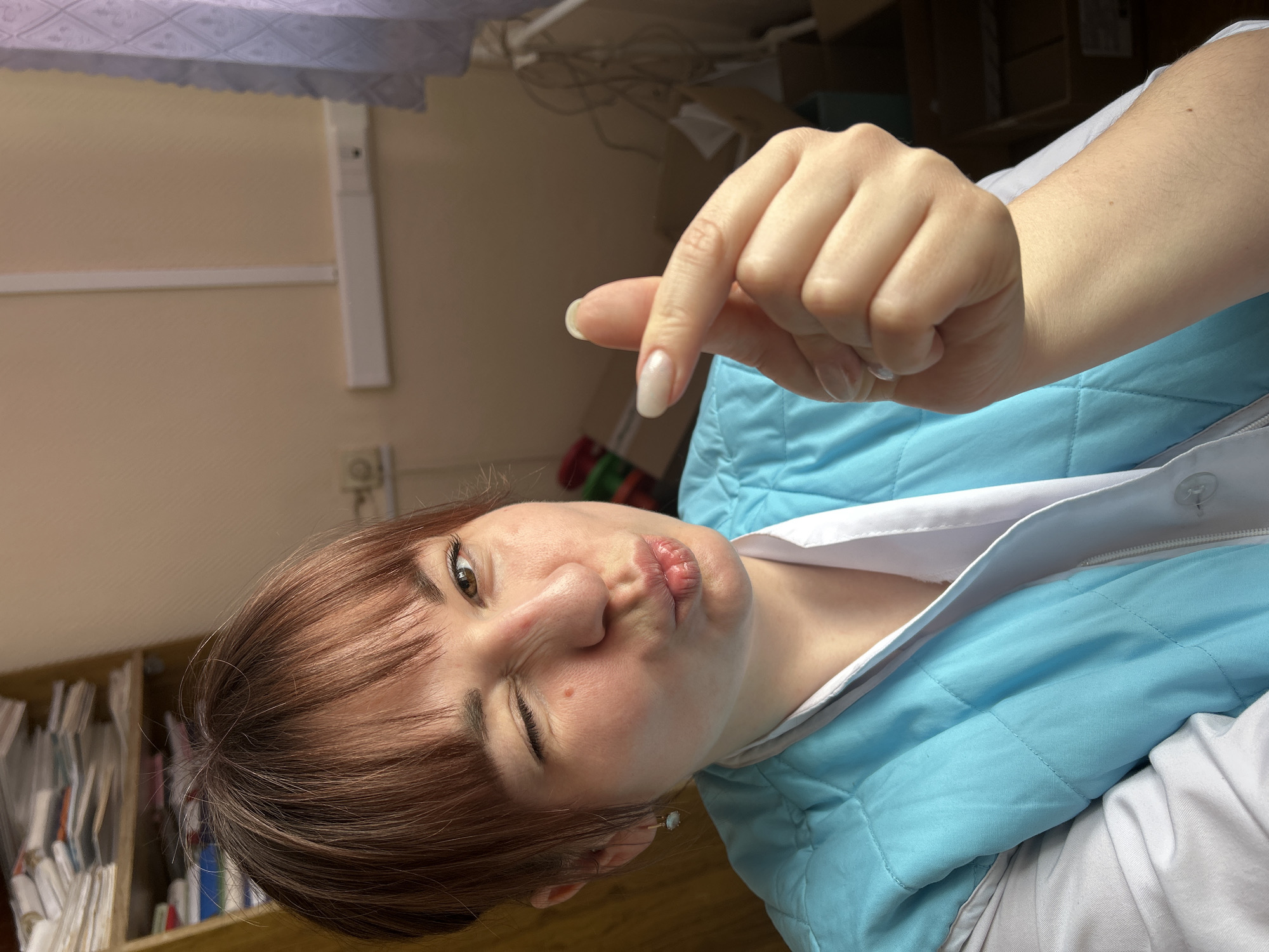 A female individual is depicted indoors in what appears to be a medical or office environment within the city of Фоки, Russia. She has light brown hair with bangs, brown eyes, and is winking her left eye while pouting her lips. She wears a light blue quilted vest over a white uniform-style top and a gold-colored hoop earring with an opalescent stone on her left ear. Her right hand is raised, forming a finger heart gesture with her thumb and index finger, showcasing long, oval-shaped nails with a light polish. The background to the upper left features a cluttered wooden shelf stacked with numerous papers, files, and books. The wall is light-colored, with white electrical conduit and an outlet visible to the right. A light-colored curtain with a diamond pattern is partially visible on the far right, and several cardboard boxes are present in the mid-ground right.