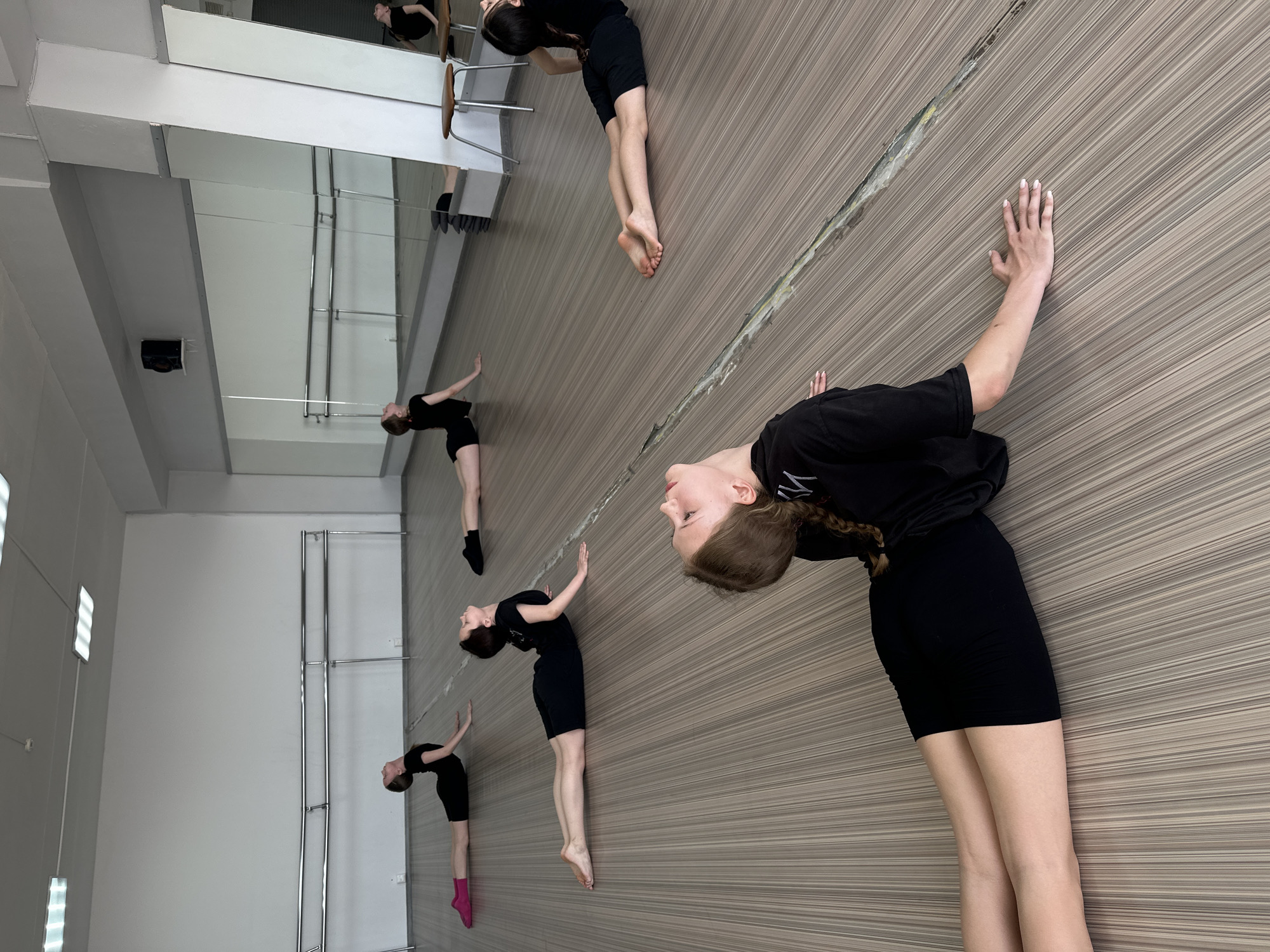 In what appears to be a dance or gymnastics studio in Votkinsk, Russia, multiple young girls are seen practicing a stretching exercise along a wall with horizontal brown lines. The girls are dressed in similar black athletic wear. They are leaning backward, with their hands placed against the wall for support. The room also includes mirrors. The floor appears to be made of linoleum.