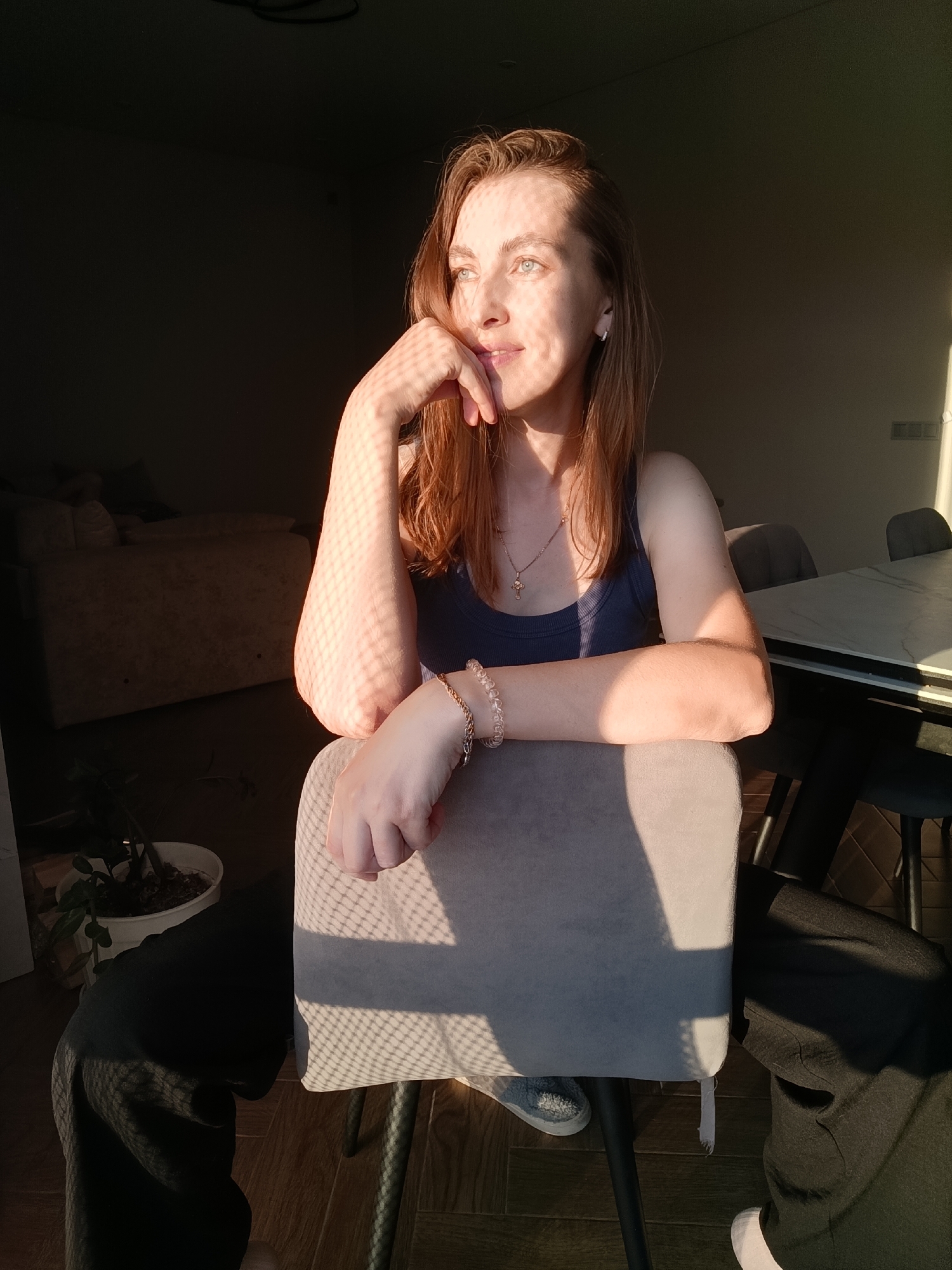 A woman with reddish-brown hair is seated in a chair, with her left elbow resting on the back of the chair and her right hand supporting her chin. She is looking off to the left, out of frame, with a contemplative expression. She is wearing a dark blue tank top and dark pants. A light-colored bracelet with clear beads is visible on her left wrist. Sunlight streams in from the right, casting a patterned shadow across her face and arm. The background is dimly lit, with a sofa visible on the left and a table and chairs on the right. The floor is made of wooden planks. The scene appears to be indoors, possibly a residential setting, in Chaikovsky, Russia.