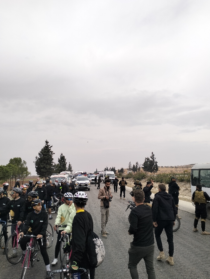 A gathering on a roadway under an overcast sky. Cyclists with bikes are grouped on the left, some wearing matching attire. Emergency vehicles, including ambulances and police cars, are positioned behind them. Individuals, some with visible tactical gear, stand on the right side of the road, near a white bus. The scene suggests a planned event, possibly a ride supported by emergency and security personnel. Trees and sparse vegetation define the landscape in the background.