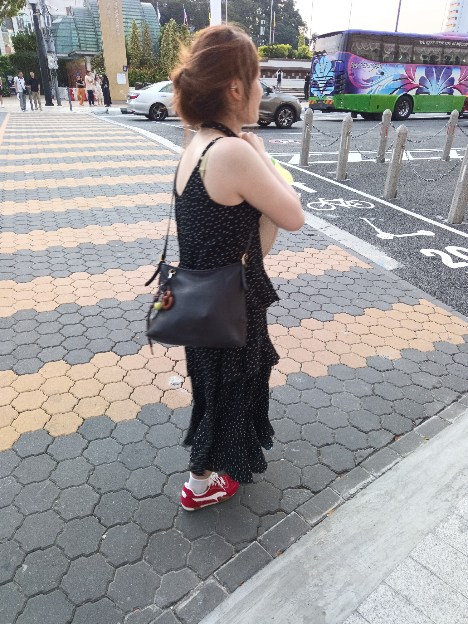 The image shows a woman walking on a sidewalk in Kuala Lumpur, Malaysia. She is wearing a black polka dot dress, white socks, and red sneakers. She is carrying a black purse over her shoulder and a yellow object in her left arm. The sidewalk is made of hexagonal paving stones in gray and yellow colors. In the background, a bus with colorful graphics and text in Malay is visible, along with a silver car and other pedestrians. The scene appears to be a street or public area in the city.