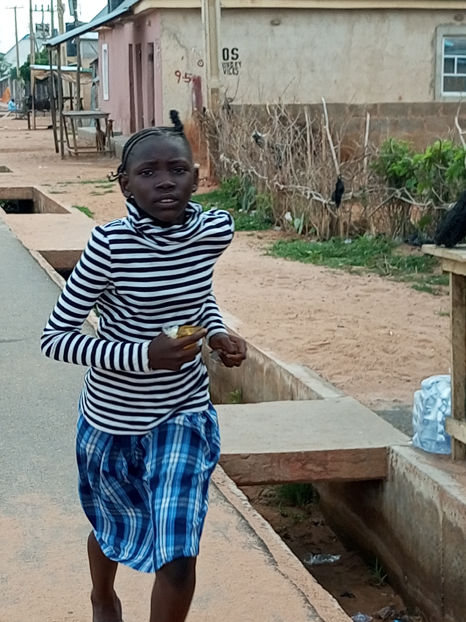 A young girl with braided hair, wearing a black and white striped turtleneck shirt and a blue checkered skirt, is standing on a paved surface next to a drainage ditch. She is holding a small object, possibly food, in her right hand and looking towards the camera with an open mouth. In the background, there are buildings, vegetation, and a street with a few indistinct figures. The environment appears to be an outdoor urban setting in the city of Gombe, Nigeria.