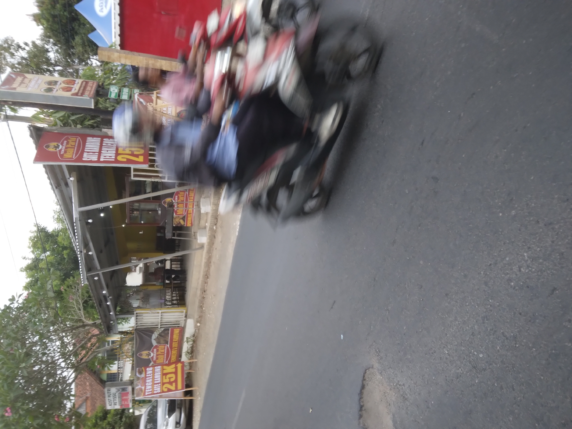 The image depicts a street scene in Desa Pamijen, Kecamatan Sokaraja, Indonesia, featuring a roadside food stall with a yellow awning and seating area. Several signs are visible, including one for "TENGKLENG SATE KAMBING MBAH PARTO" with a price of "25K". In the foreground, two individuals on a motorcycle are captured in motion, creating a blur effect. A white car is parked on the left side of the road. The environment includes a paved road with some patches and greenery in the background, suggesting a semi-rural or suburban setting.