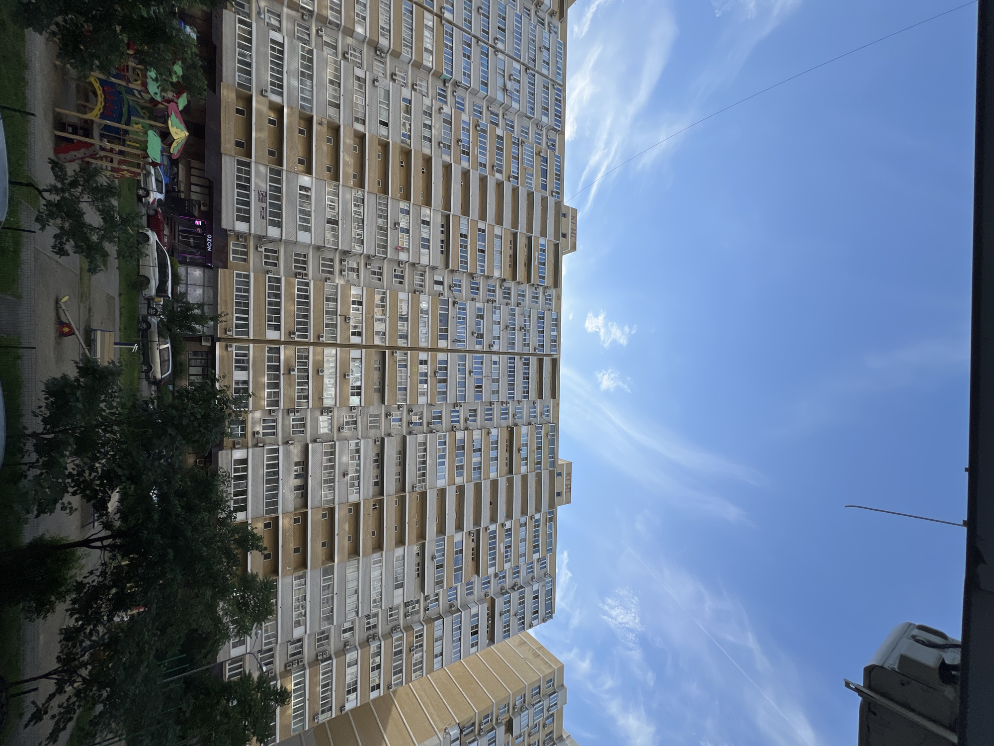The image, oriented in portrait, displays a towering residential apartment building under a bright, clear blue sky. The multi-story structure occupies the left side of the frame, featuring a repetitive pattern of windows and enclosed balconies, adorned with numerous external air conditioning units. The building's facade alternates between light grey brick-like sections and beige panels.
At ground level, mature trees line a paved area, partially obscuring a colorful children's playground. Several cars are parked along what appears to be a street or parking lot adjacent to the building. Commercial establishments are visible on the ground floor, with a prominent "OZON" sign clearly displayed on one of them. A portion of another, shorter apartment building can be seen in the lower right foreground.
The right side of the image is dominated by the expansive blue sky, dotted with scattered, wispy white clouds, indicating a sunny daytime. A thin, dark wire stretches horizontally across the sky. Faint Cyrillic lettering, possibly "КИРИ," is discernible on an upper facade section of the main building. The scene depicts a typical urban residential environment in bright daylight.