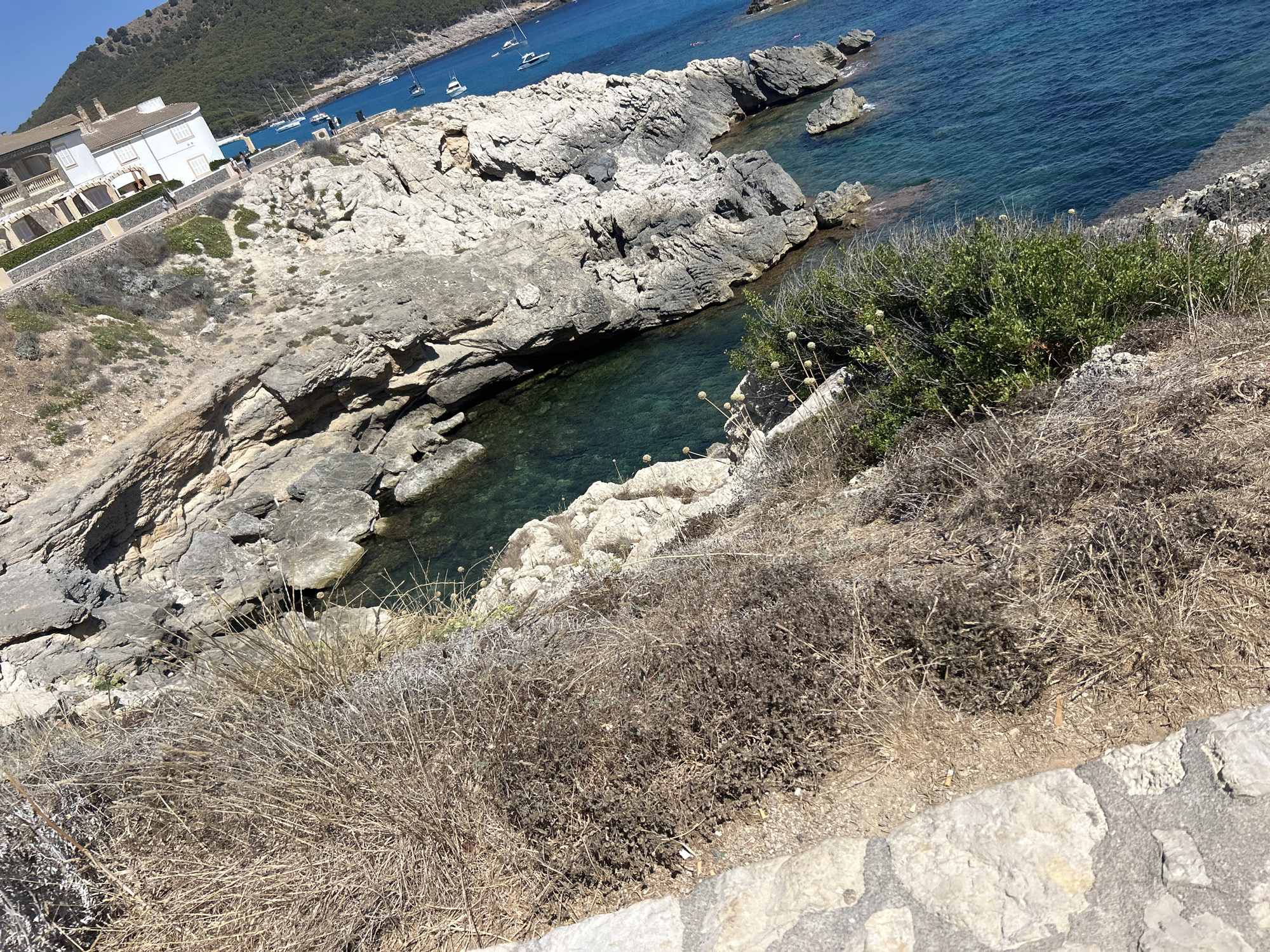 The image shows a scenic view of Cala Lliteres, Spain, featuring rugged coastal terrain overlooking a turquoise bay. A white building complex with red-tiled roofs is visible on the left, situated on a hillside. Several sailboats are anchored in the clear blue water. The foreground is dominated by dry, sun-baked vegetation and rocky outcrops, indicative of a Mediterranean climate. The water is clear, revealing the rocky seabed in shallow areas. The scene is bathed in bright sunlight, suggesting a clear day.