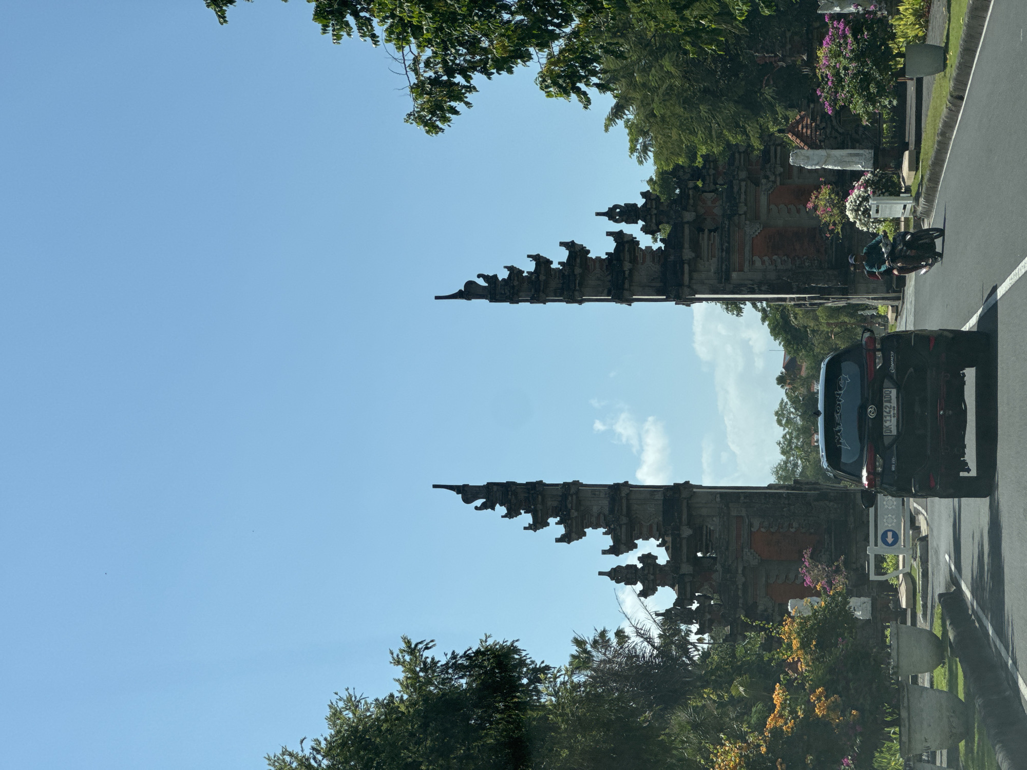 A black SUV drives on a paved road in Kuta Selatan, Indonesia. The SUV is in the foreground of the image, with its rear facing the camera. To the left of the road, there are large planters filled with green grass and orange flowers. To the right of the road, there is a motorcycle with two people riding on it. In the background, there is a large, ornate gate with two towers. The gate is made of stone and has intricate carvings. The sky is clear and blue, with a few white clouds. The scene appears to be a sunny day, likely in the afternoon.