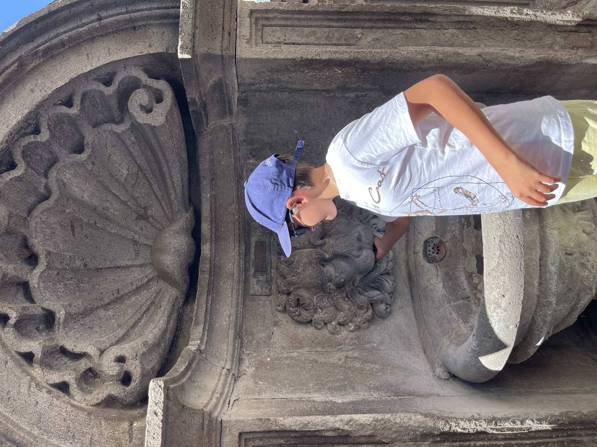 A young boy wearing a blue baseball cap, sunglasses, a white t-shirt, and yellow shorts stands in front of a stone fountain in Viterbo, Italy. The fountain is ornately carved with a shell motif at the top and a lion's head, from which water appears to be flowing into the basin below. The boy is reaching out to touch the lion's head. The stone structure appears old and weathered.
