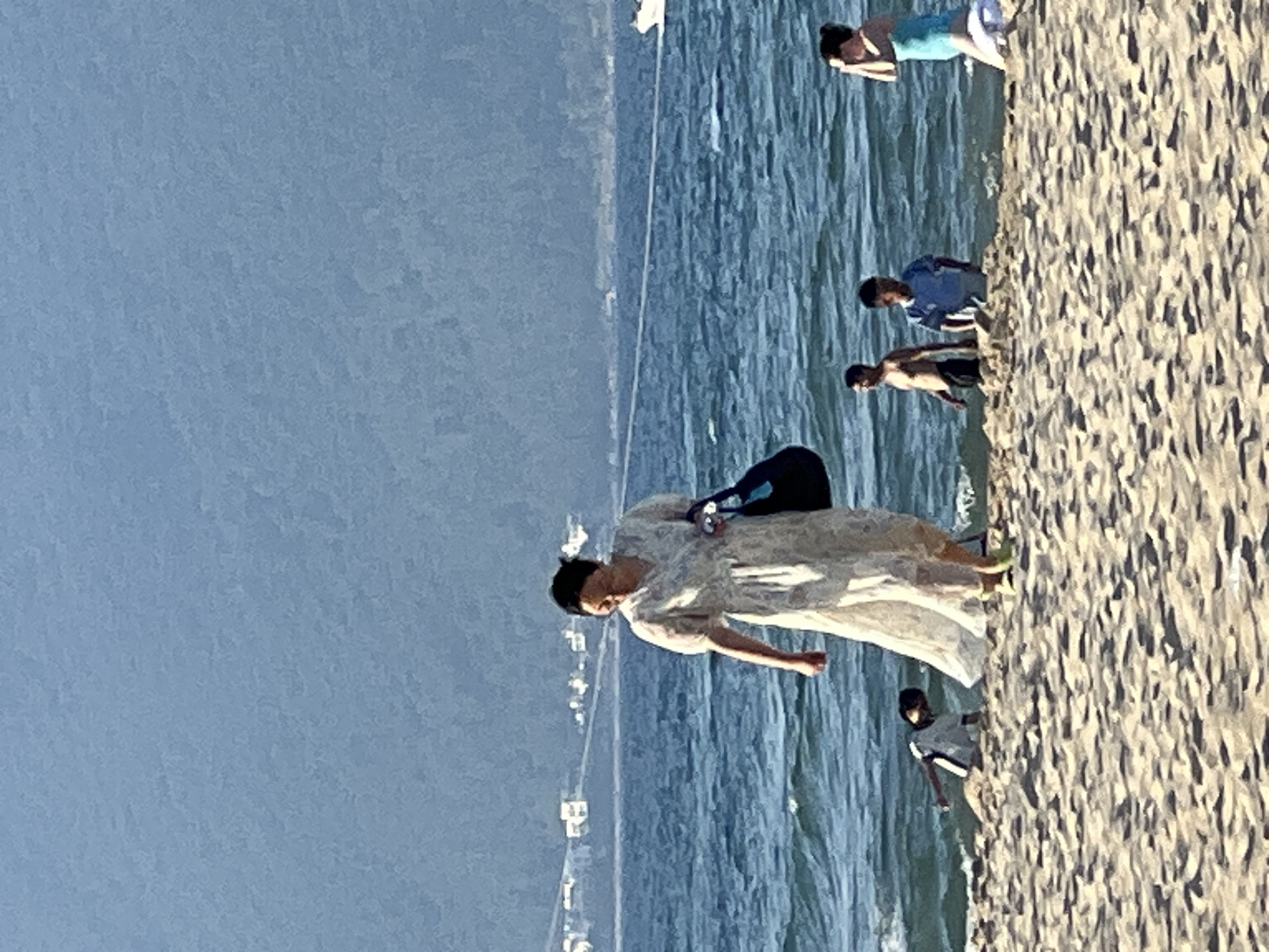 This image captures a sunny day at a beach in Da Nang, Vietnam. The scene depicts several people near the water's edge and on the sandy shore. In the foreground, a person in a light-colored dress is bent over, with their arms extended. To their left, a person in a blue bathing suit is partially visible. Further back, a group of people are gathered near the water. One person in a blue shirt is looking down, while another person, shirtless, stands beside them. In the distance, more individuals are visible, some wading in the ocean. The water is a vibrant blue, with gentle waves rolling onto the shore. The beach is covered in light-colored sand, with some areas appearing wet from the tide. The bright sunlight suggests it is likely midday or early afternoon. The overall atmosphere appears relaxed and leisurely, with people enjoying a day at the beach. There is no visible text in the image.