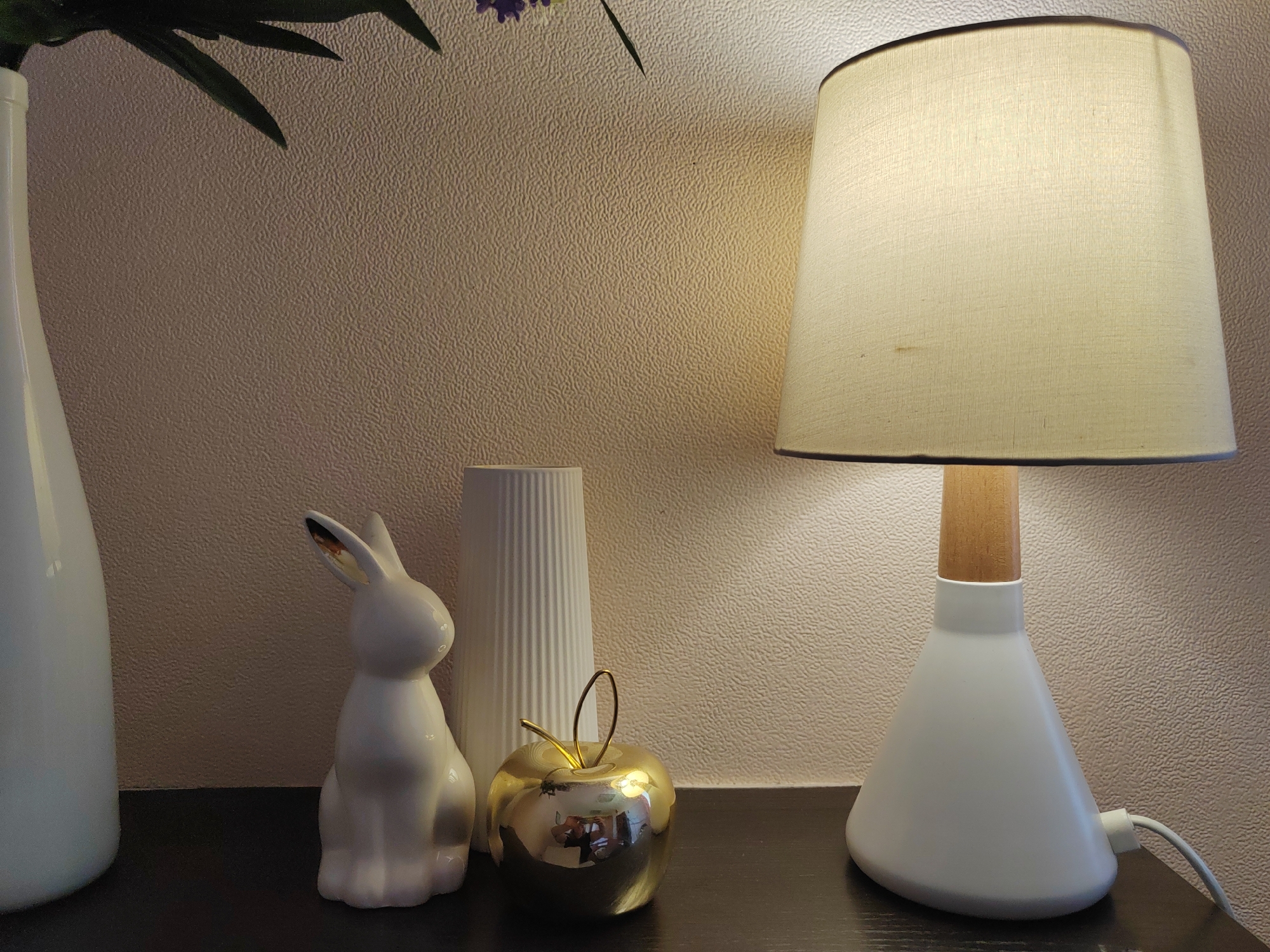 The image displays a still life arrangement on a dark surface against a textured, light-colored wall. From left to right, a tall, white, ceramic vase with a smooth finish is visible. Next to it sits a white, ceramic rabbit figurine, its ears perked forward. Behind the rabbit is a tall, white, ribbed vase or cylindrical object. In front of the ribbed vase and to the right of the rabbit is a small, gold, apple-shaped decorative object with a gold wire stem. To the far right is a white table lamp with a conical base and a light-colored fabric lampshade, emitting a warm glow. The scene is indoors, likely in a home or studio setting, with no indication of a specific city or location.