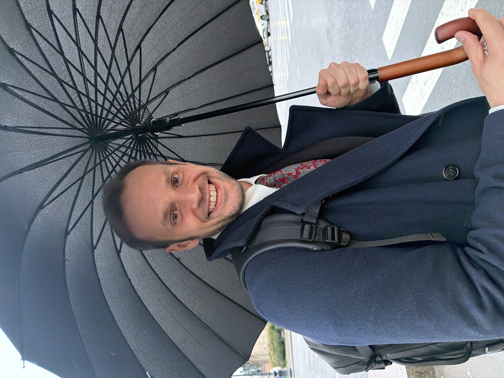 The image shows a man holding a large, black umbrella with a wooden handle. He is smiling and wearing a dark blue overcoat over a white shirt and patterned tie. He also has a backpack on. The scene is set on a street in Saint Petersburg, Russia, as evidenced by the street markings and typical city elements visible in the background. The weather appears to be wet or rainy. There are crosswalk markings on the street.