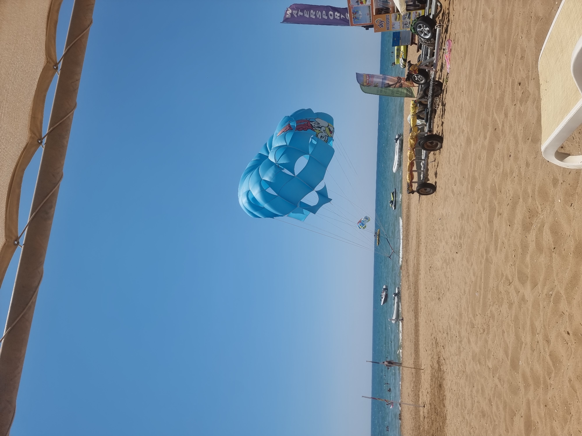 An aerial view captures a bright blue sky above a sandy beach bordering a calm, blue ocean. A large, bright blue parachute with a cartoon character design is visible in the sky, attached to a boat that is out of frame. The beach is lined with a few palm trees and some beach chairs, and there are several jet skis and boats in the water. The scene appears to be a typical beach resort area, likely in Manavgat, Turkey, given the presence of watersports equipment and the general atmosphere of a tourist destination. There are a few people scattered on the beach and in the water, enjoying the sunny weather. A trailer with yellow fabric is parked on the sand, and several promotional banners are set up near the beach. The overall impression is a vibrant and active beach scene on a clear, sunny day.