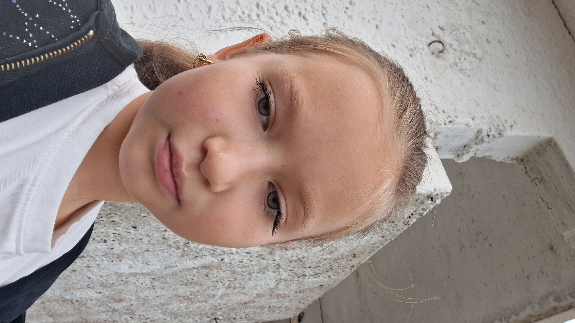 The media file is a close-up, tilted, portrait-oriented photograph of a young girl. She has fair skin, light brown hair, and green eyes with noticeable mascara. She is wearing a white t-shirt and a dark jacket with a zipper. A small golden earring is visible in her left ear. The background is a textured, light-colored surface, possibly concrete, with some debris. The lighting suggests it might be daytime, outdoors. The girl's expression is neutral, with slightly parted lips. The image appears to be a candid shot, possibly a selfie, given the angle. There is no visible text in the image. The setting suggests an outdoor urban or industrial environment.