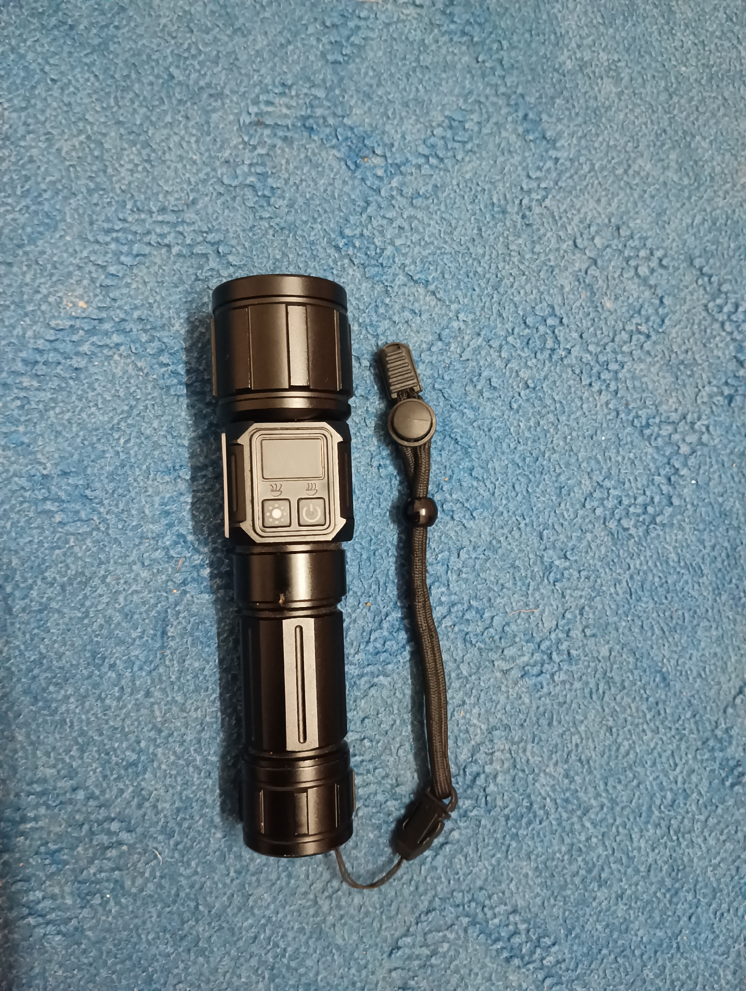 A black flashlight with a digital display and two buttons lies on a textured blue surface, possibly a rug or towel. A black lanyard with a toggle and bead is attached to the flashlight. The flashlight is positioned vertically, slightly to the left of the center of the image. The blue surface has a soft, fuzzy texture and appears to be a solid color. There are no people visible in the image, and no identifiable landmarks or specific location details, including Manali, India, are present or suggested. The lighting is even, and the image is in focus.