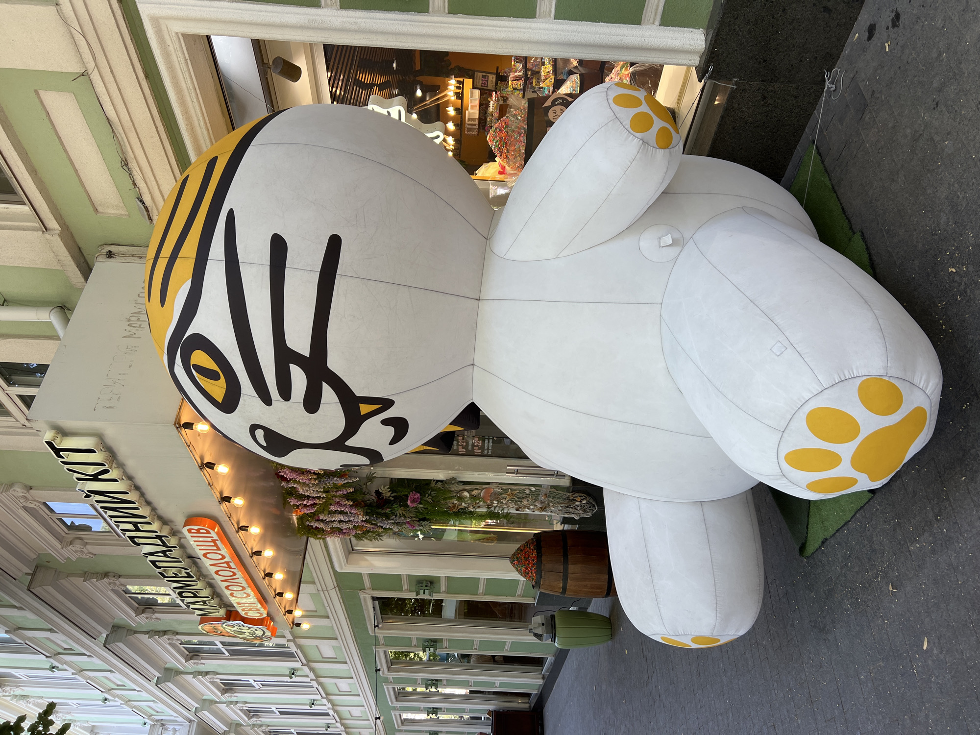 An inflatable, cartoonish tiger character is displayed prominently outside a store with a green facade. The tiger is predominantly white with yellow and black markings, including stripes on its head and paw prints on its feet. The store has a sign in Cyrillic script that reads "МАРМЕЛАДНИЙ КІТ" (Marmeladnyy Kit), which translates to "Marmalade Cat". Below this, another sign reads "СВІТ СОЛОДОЩІВ" (Svit Solodoshchiv), meaning "World of Sweets". The store appears to be a candy shop, with colorful sweets visible through the display window. Decorative garlands of flowers and seashells hang near the entrance. To the left of the inflatable tiger, there is a wooden barrel filled with small red flowers and a tall, green, cylindrical trash can. The scene is set on a paved sidewalk in front of a building with classical architectural elements. The image was taken in Odesa, Ukraine. There are no people visible in the scene.